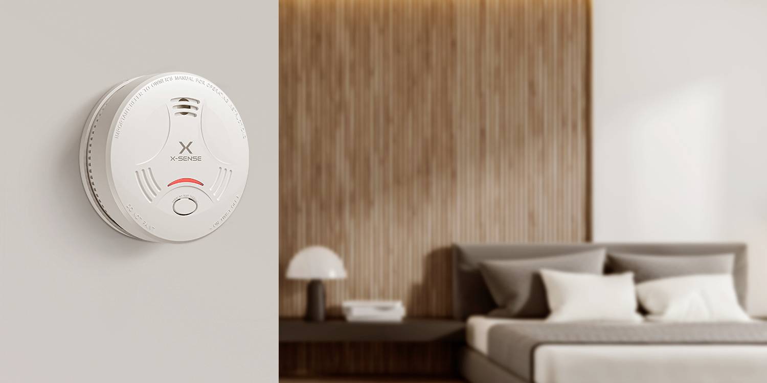 Ein weißer Rauchmelder mit blinkendem rotem Licht ist an einer Wand in einem modernen Schlafzimmer mit einem ordentlich gemachten Bett und einem Nachttisch montiert.