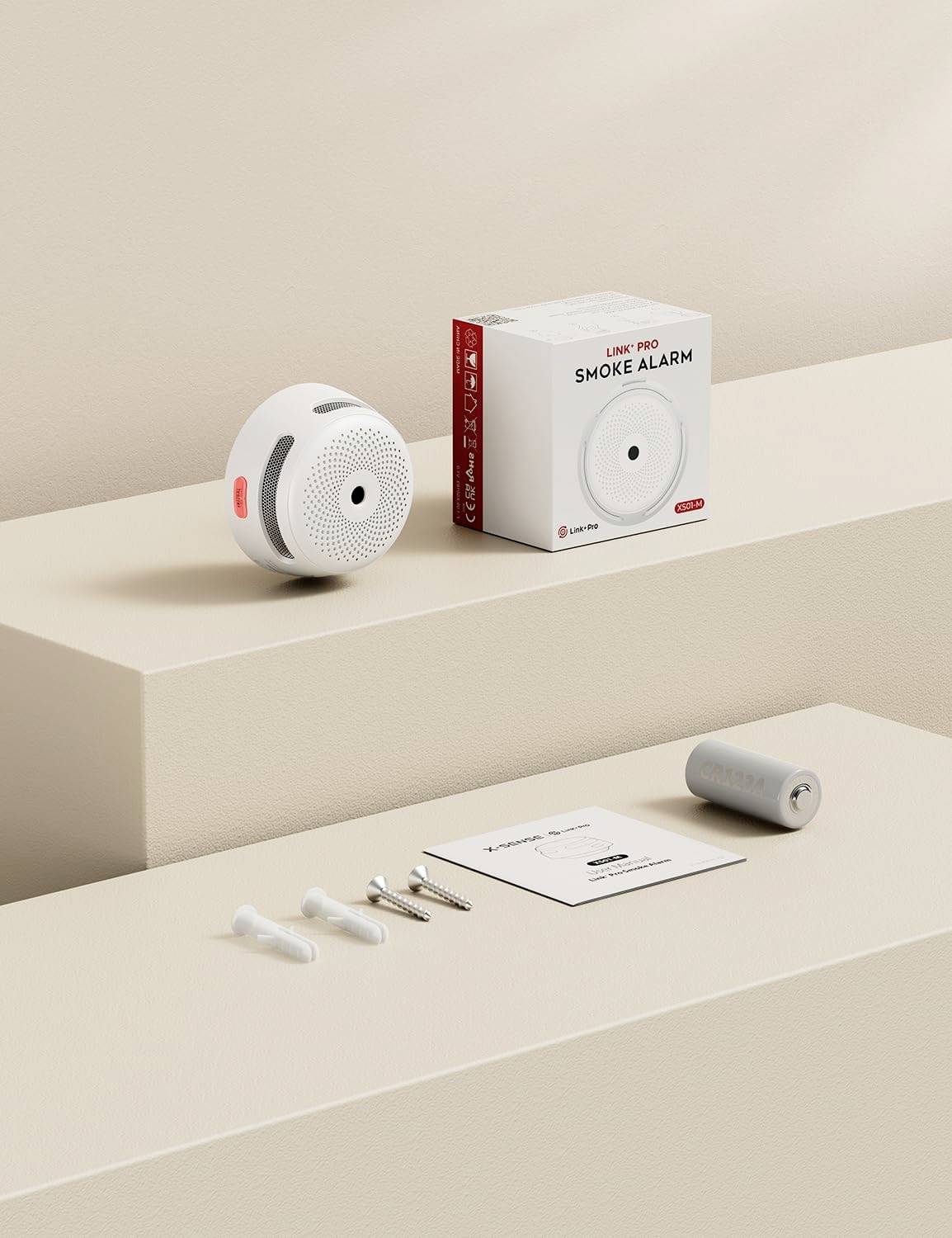 Eine Rauchmeldereinheit mit kreisförmigem Design, eine Verpackungsbox, Befestigungsmaterial und eine Bedienungsanleitung werden auf beigen Plattformen präsentiert.