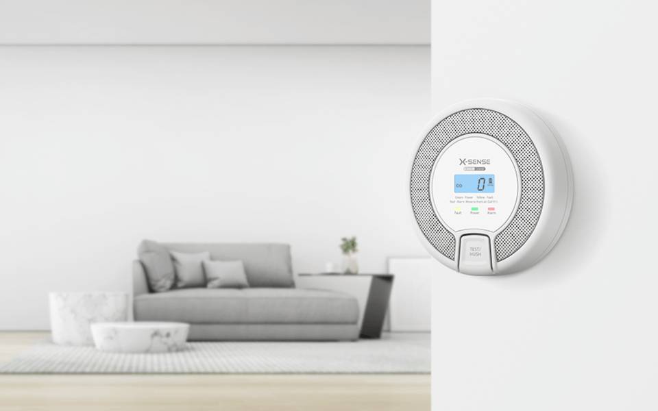 Ein Rauch- und Kohlenmonoxidmelder ist an einer weißen Wand montiert. Im Hintergrund ein modernes Wohnzimmer mit einem grauen Sofa und Dekorationen.