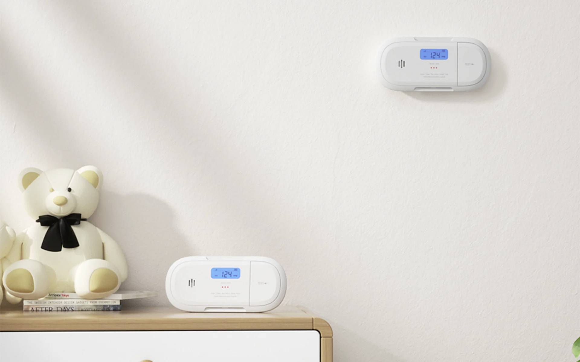 Ein Raum mit einem weißen Teddybären auf einer Kommode neben einem Rauch- und Kohlenmonoxidmelder. Ein weiterer ähnlicher Melder ist an der Wand befestigt.