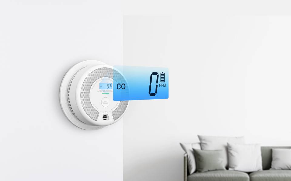 Ein digitaler Kohlenmonoxid-Detektor an einer weißen Wand zeigt eine Anzeige von "0 PPM". Ein grauer Couchtisch ist im Hintergrund teilweise zu sehen.