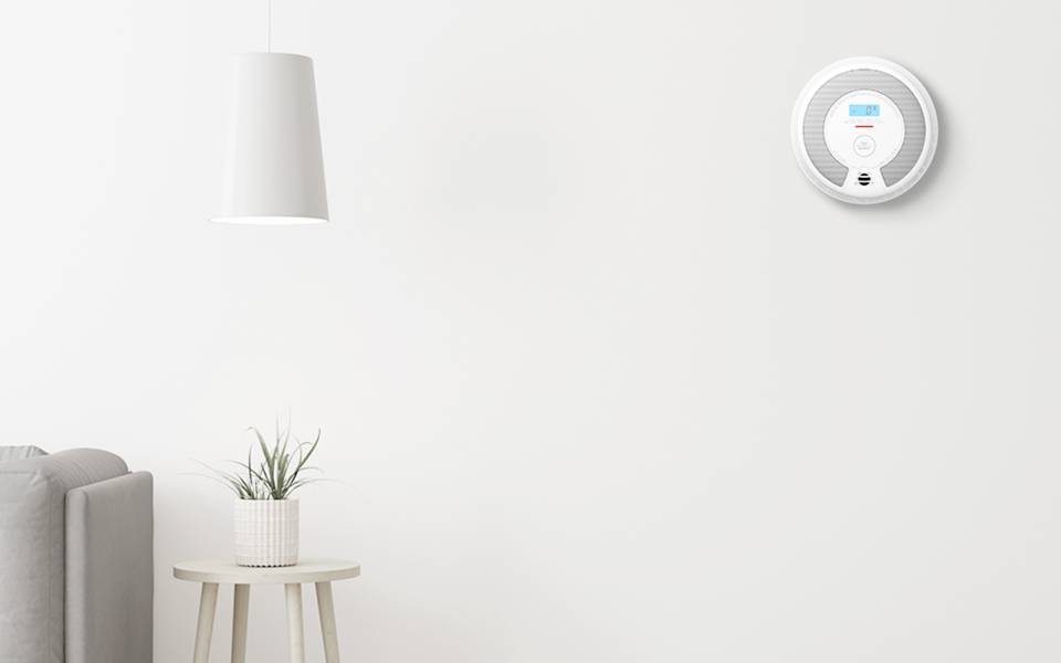 Ein moderner Wohnraum mit einer weißen Wand zeigt einen intelligenten Thermostat, der neben einer Hängelampe und einer Pflanze auf einem kleinen Tisch die Temperatur anzeigt.