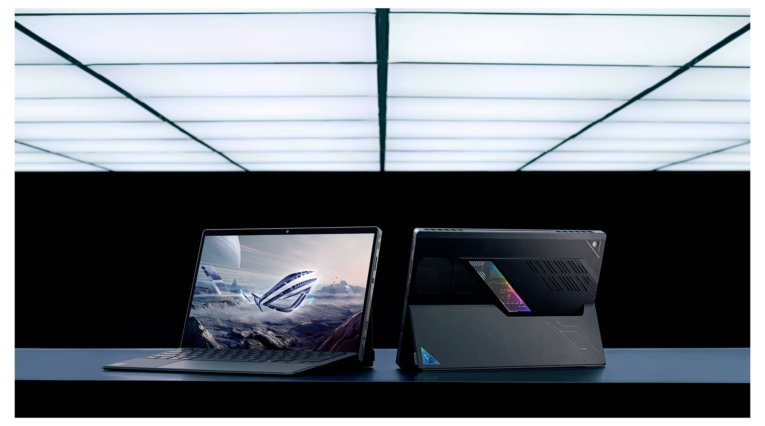 Zwei Laptops stehen in verschiedene Richtungen auf einem dunklen Tisch unter hellen, segmentierten Deckenleuchten. Auf einem Bildschirm werden futuristische Grafiken angezeigt.