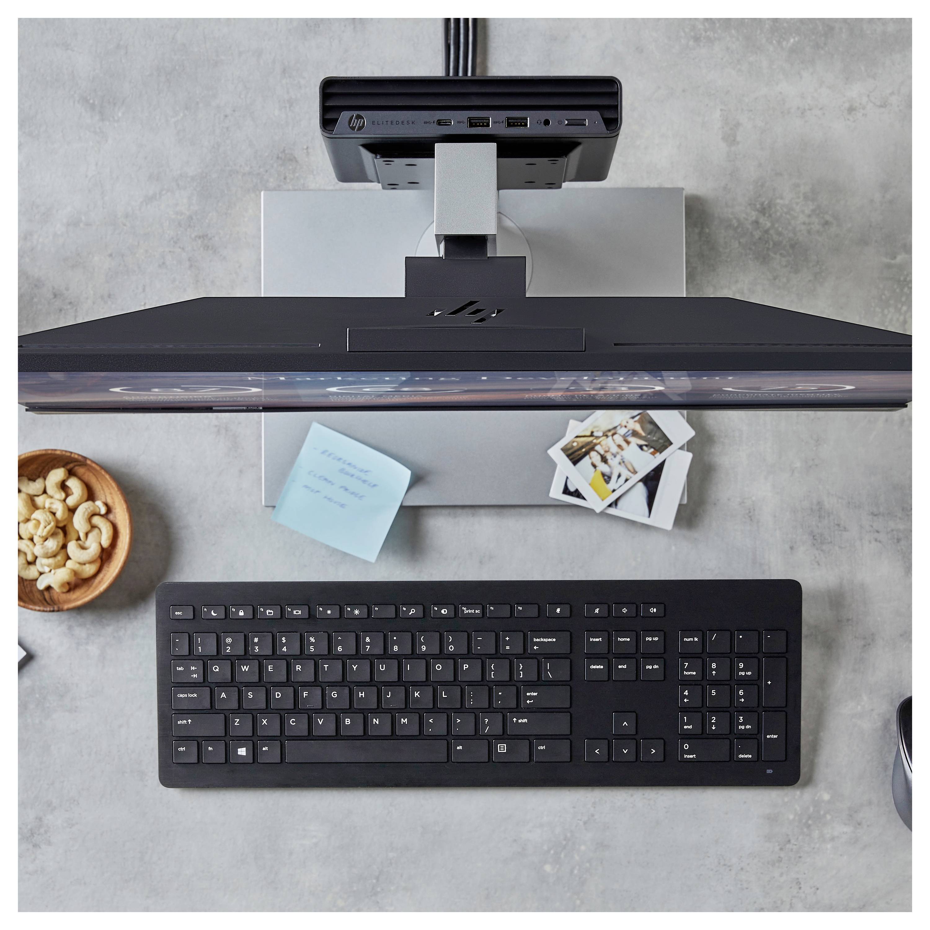 Eine Draufsicht eines Arbeitsplatzes mit einer schwarzen Tastatur, einem Monitor, Haftnotizen, Cashewnüssen in einer Schale und Fotos auf einem grauen Tisch.