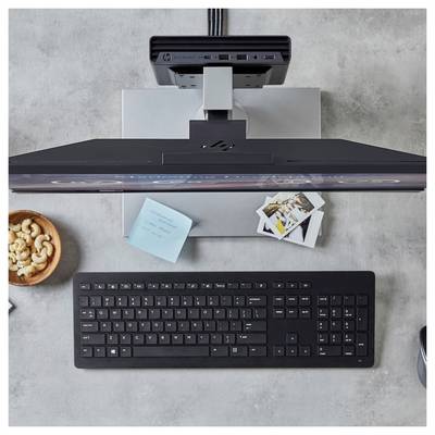 Eine Draufsicht eines Arbeitsplatzes mit einer schwarzen Tastatur, einem Monitor, Haftnotizen, Cashewnüssen in einer Schale und Fotos auf einem grauen Tisch.
