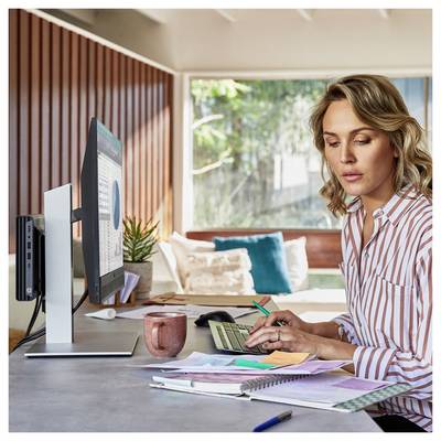 Eine Frau, die an einem Schreibtisch mit einem Computer arbeitet und einen Stift über Papiere hält. Sonnenlicht durchflutet den Raum, in dem im Hintergrund eine Couch steht.