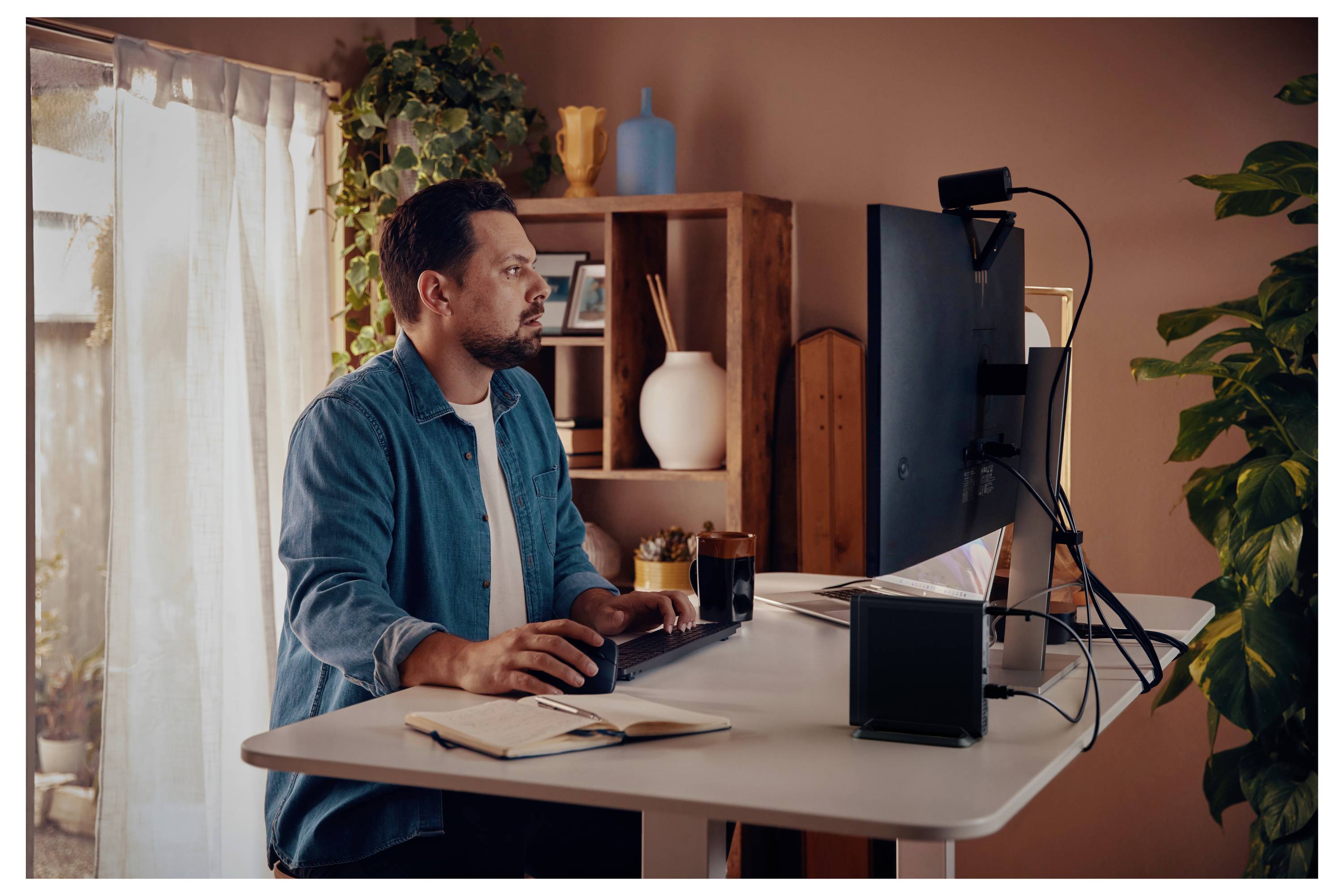 Ein Mann konzentriert sich intensiv auf eine Aufgabe und arbeitet an einem Computer in einem Heimarbeitsplatz. Ein Notizbuch und ein Getränk befinden sich in der Nähe.