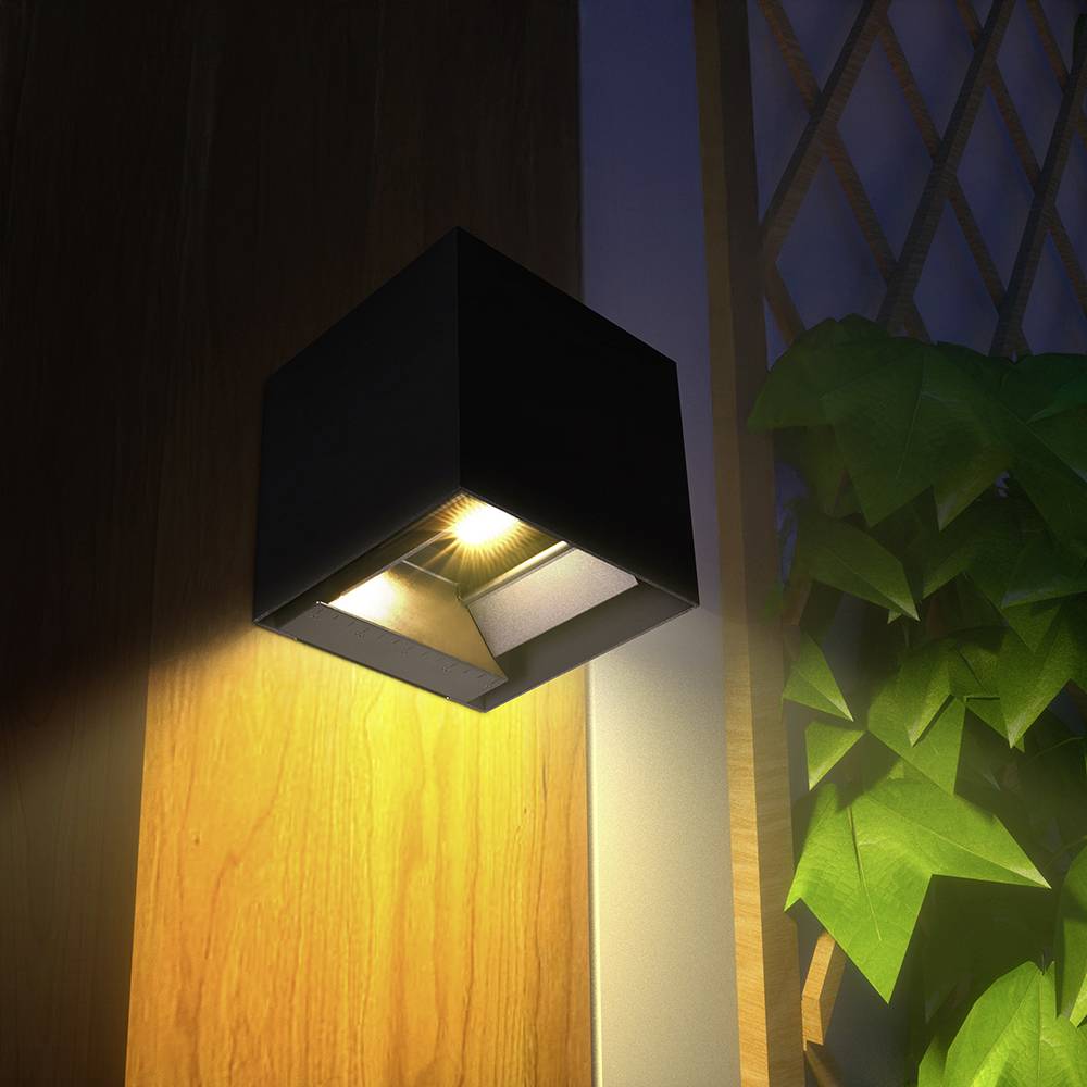 Eine moderne wandmontierte Leuchte mit quadratischem Design beleuchtet eine Holzvertäfelung und wirft weiches Licht auf die umgebende Vegetation.