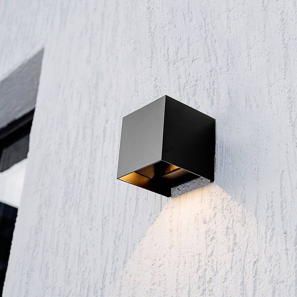 Eine schlichte schwarze, würfelförmige Wandleuchte strahlt weiches Licht vor einer strukturierten grauen Wand aus und erzeugt eine moderne, minimalistische Ästhetik.