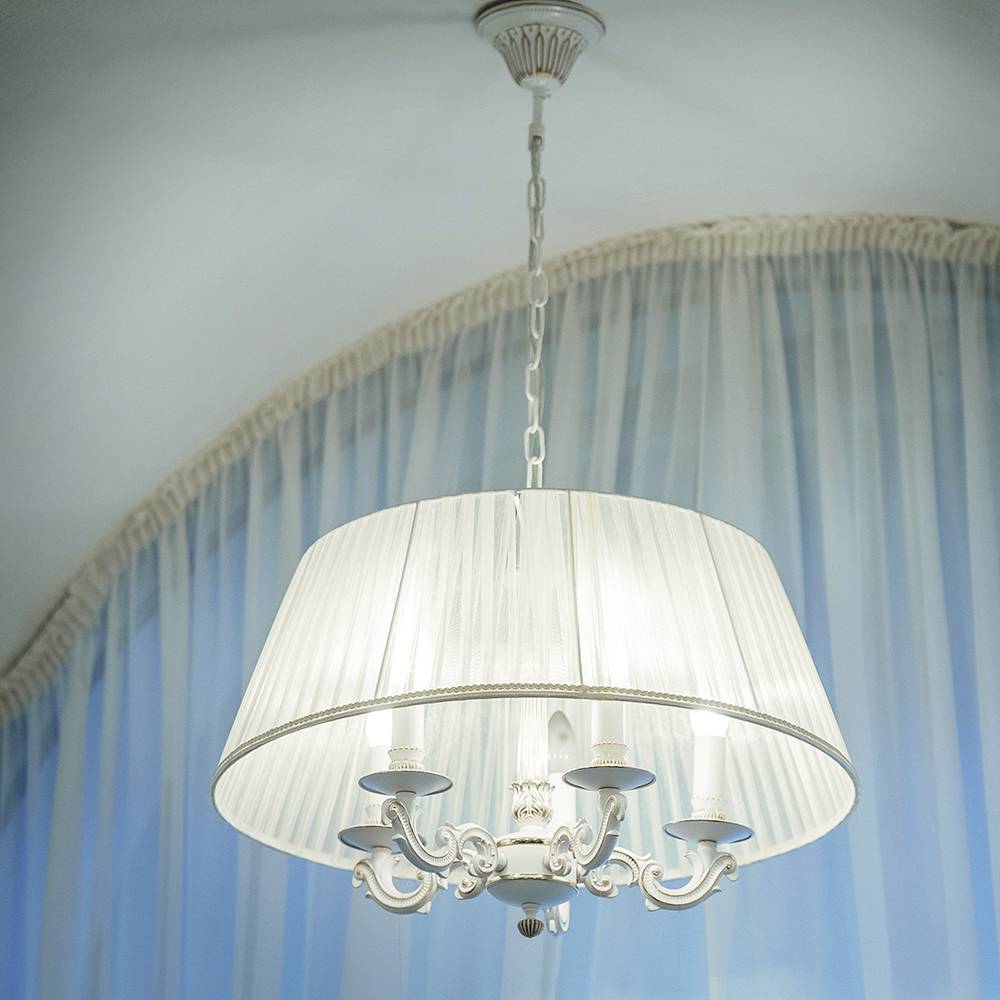 Ein hängender Kronleuchter mit einem weißen Stoffschirm und ornamentalem Design, sanft beleuchtet, vor transparenten blauen Vorhängen in einem eleganten Innenraum.