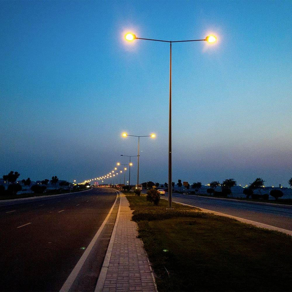 Eine friedvolle, leere Straße in der Abenddämmerung mit leuchtenden Straßenlaternen, die sich in die Ferne erstrecken, gesäumt von Grünflächen und einem blauen Himmel darüber.