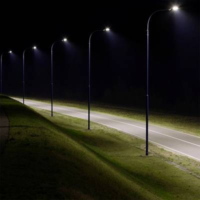 Eine Reihe von Straßenlaternen beleuchtet eine einspurige Straße bei Nacht und hebt den Grasbewuchs auf beiden Seiten vor einem dunklen Hintergrund hervor.