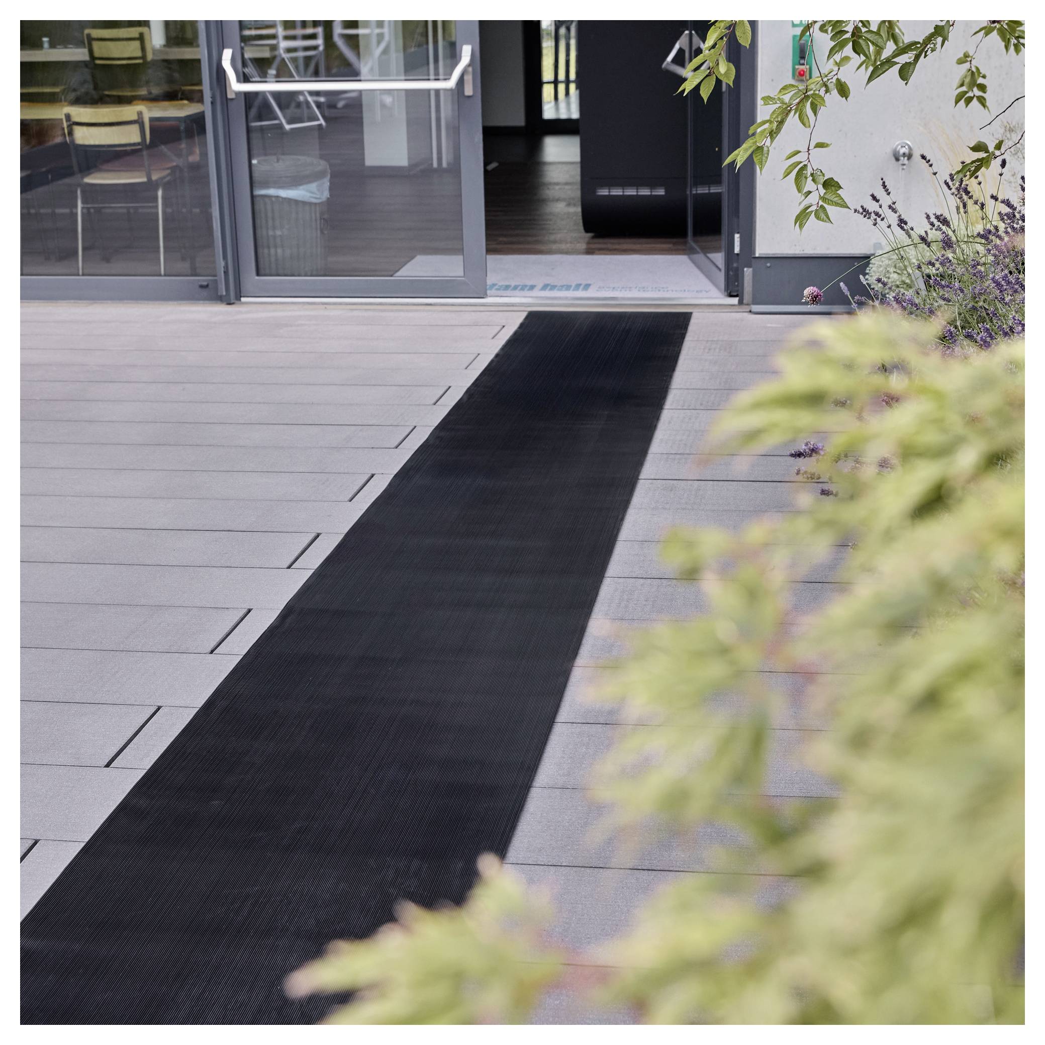 Eine schwarze Gummiraupe führt zu einem offenen Glaseingang mit sichtbaren Treppen im Inneren. In der Nähe befinden sich Außensitzplätze und grüne Pflanzen.