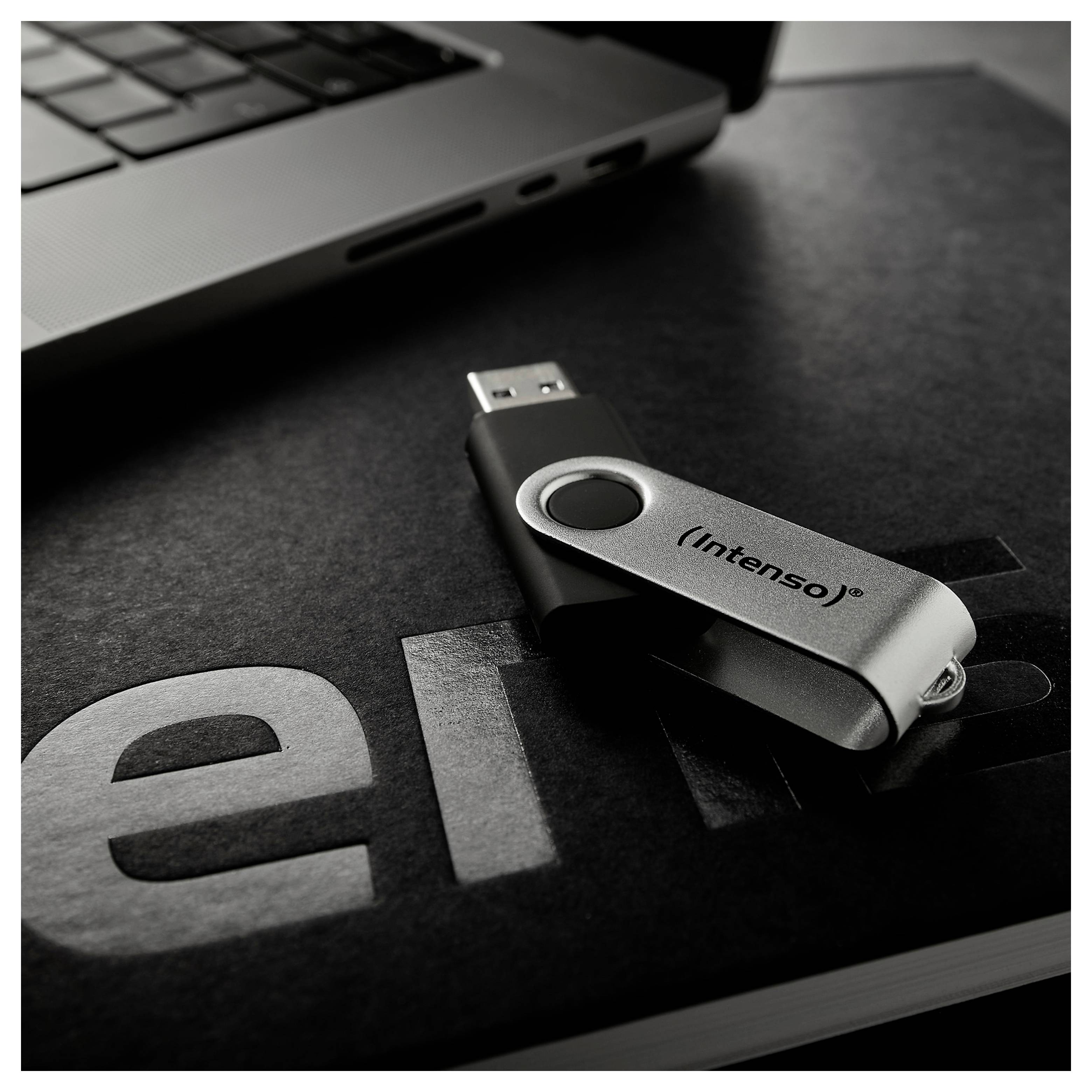 Ein silberner USB-Stick mit der Aufschrift "Intenso" liegt auf einem Notizbuch, neben einem teilweise sichtbaren Laptop, was auf Datentransfer oder Speicherung hindeutet.