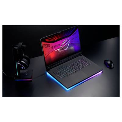 Ein Gaming-Laptop mit einer beleuchteten Tastatur zeigt das ‚ROG STRIX' Logo auf seinem Bildschirm. Kopfhörer und eine Maus sind in der Nähe platziert.