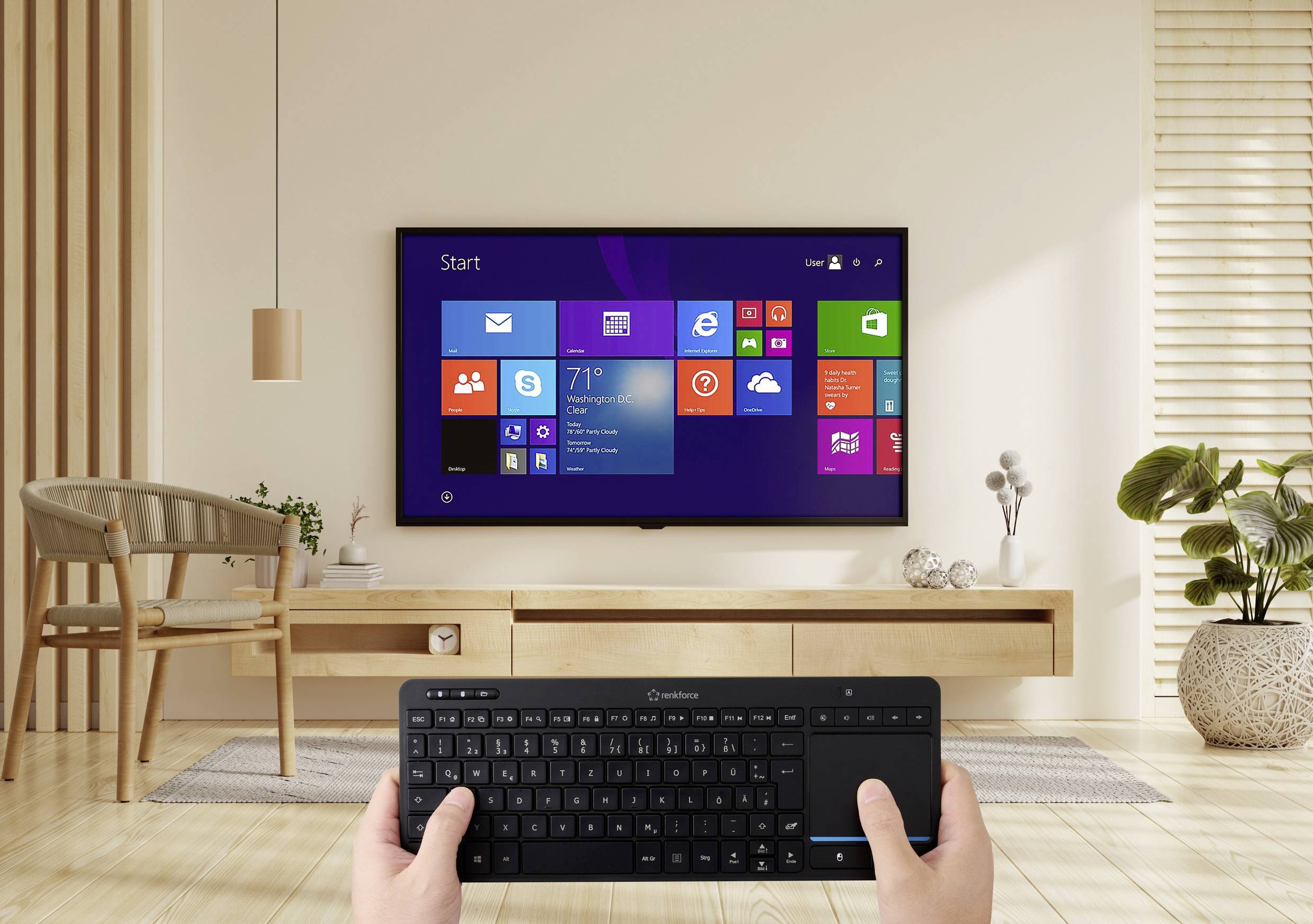 Ein Wohnzimmer mit einem an der Wand montierten Fernseher, auf dem ein Windows-Startbildschirm zu sehen ist. Eine Person hält eine kabellose Tastatur und steuert damit den Fernseher.
