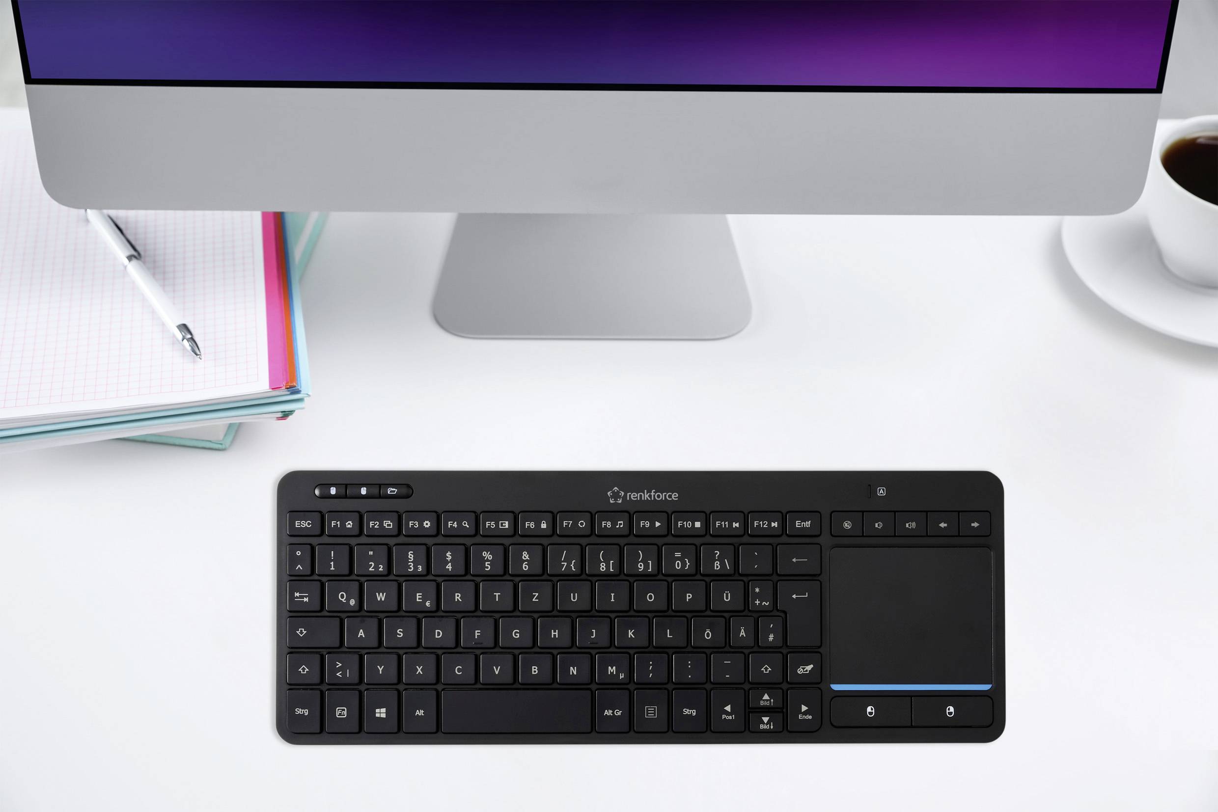 Eine kabellose Tastatur mit integriertem Touchpad liegt auf einem weißen Schreibtisch vor einem iMac. Ein Notizbuch liegt neben einer Tasse Kaffee.