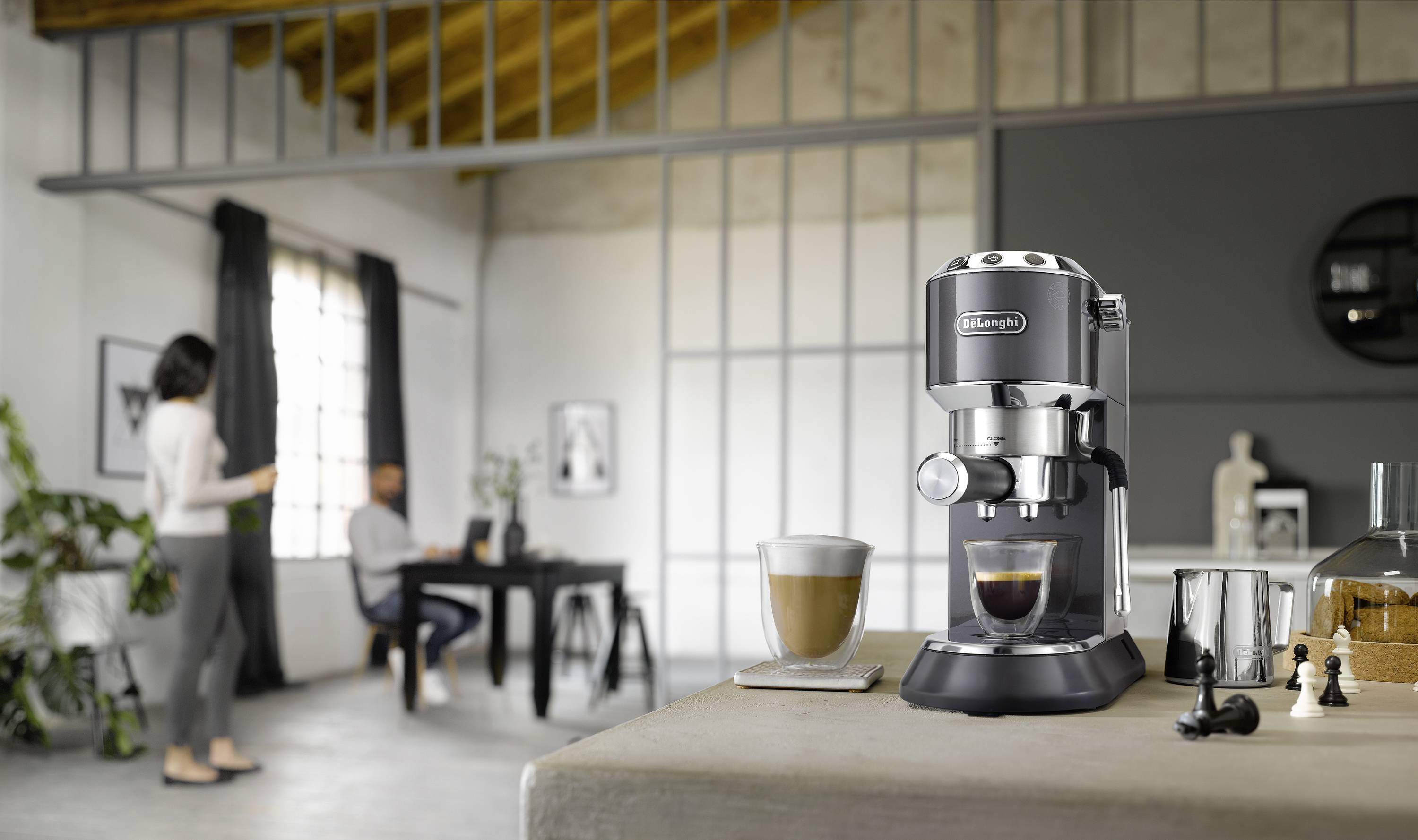 Eine elegante Espressomaschine mit einem Milchaufschäumer, die Kaffee in einer modernen Küche braut. Zwei Personen unterhalten sich im Hintergrund.