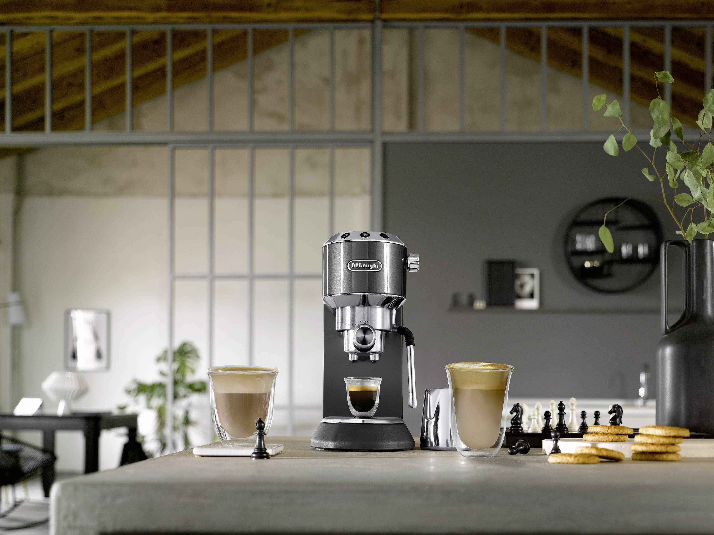 Eine kompakte Espressomaschine auf einer Küchenarbeitsplatte, umgeben von zwei Tassen Kaffee, Keksen und Schachfiguren in einem modernen Loft-Ambiente.