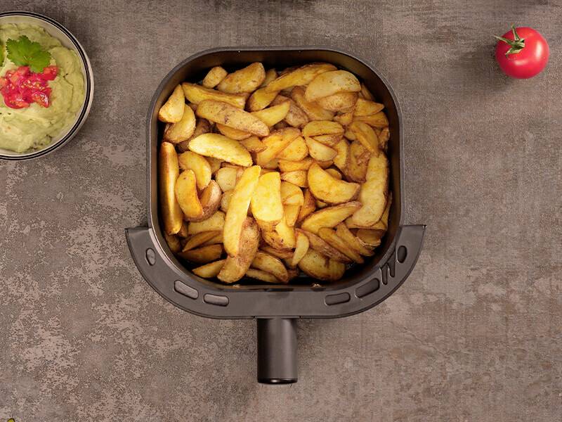 Ein Heißluftfritteuse-Korb gefüllt mit gebratenen Kartoffelecken steht auf einer Steinarbeitsplatte, begleitet von einer Schüssel Dip und einer Kirschtomate.