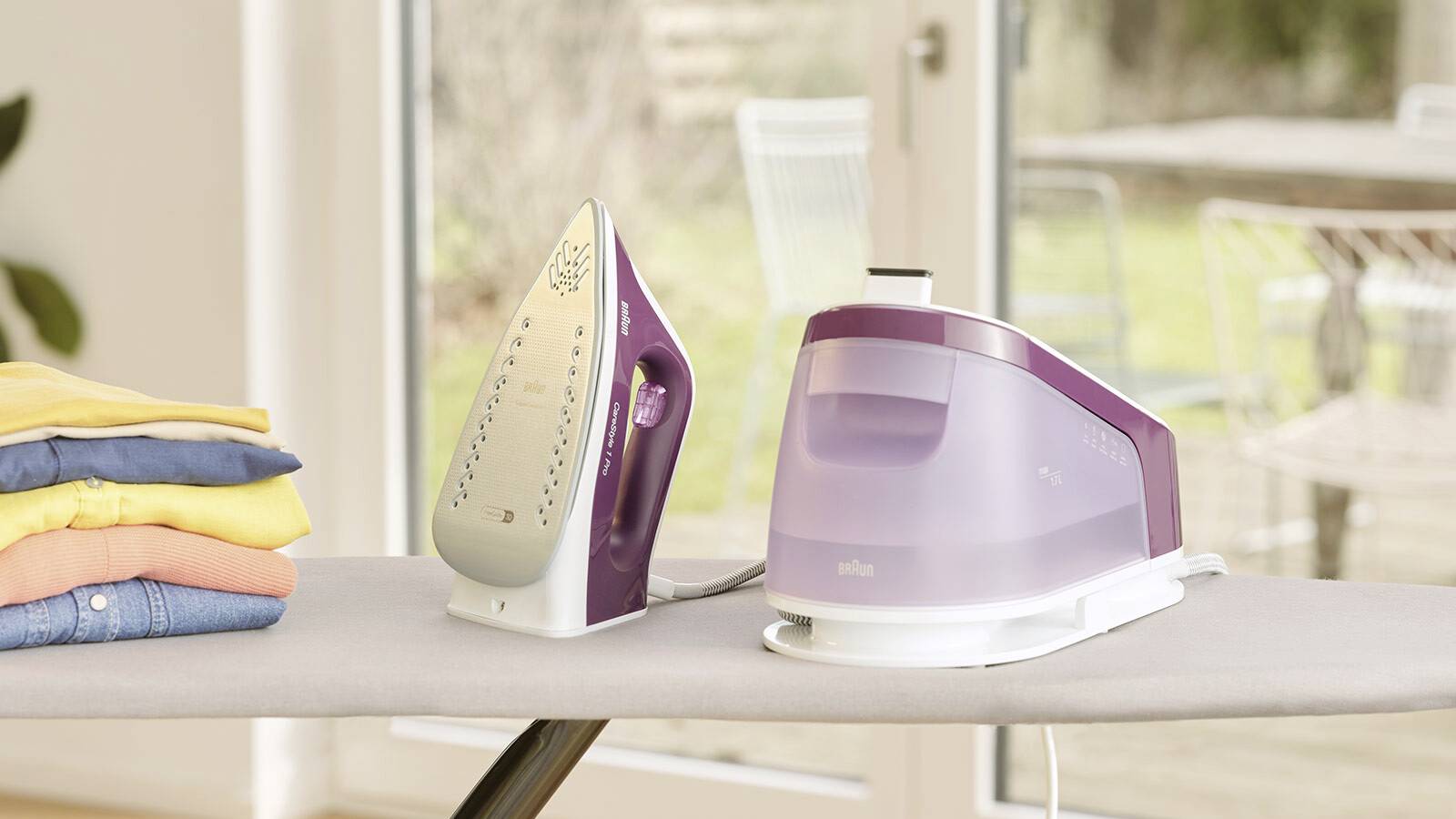 Ein lila Dampfbügeleisen und seine Basis stehen auf einem Bügelbrett neben einem gefalteten Stapel bunter Kleidung, mit einem Fenster im Hintergrund.