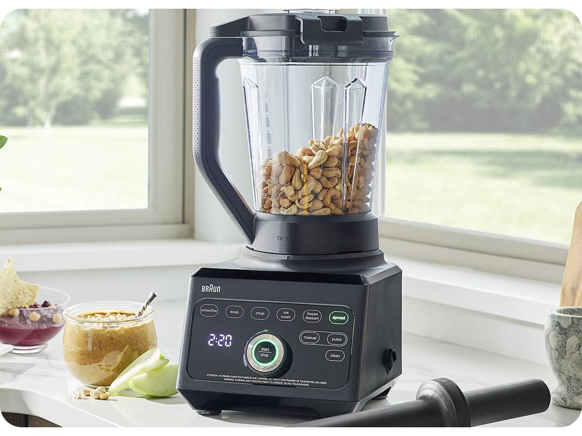 Ein Mixer auf einer Küchenarbeitsplatte, gefüllt mit Nüssen, die digitale Anzeige auf '2:20' eingestellt, neben einer Schüssel mit Dip und Tortillachips.