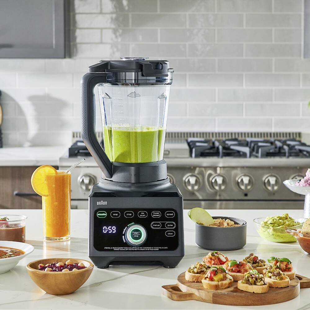 Ein moderner Mixer auf einer Küchenarbeitsplatte mixt einen grünen Smoothie, umgeben von verschiedenen zubereiteten Gerichten und einem Orangensaft.