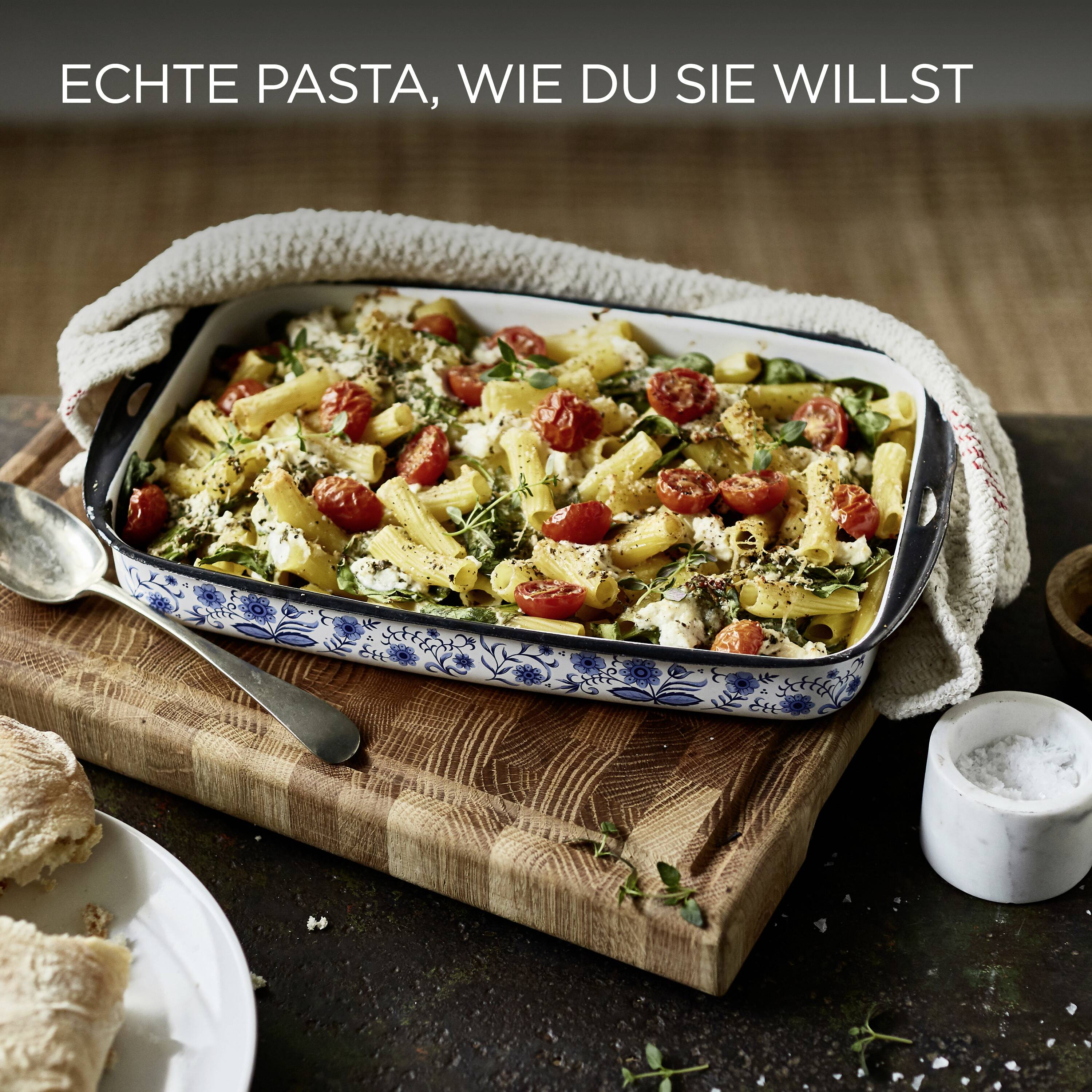 Ein gebackenes Nudelgericht mit Kirschtomaten, Käse und Kräutern in einer Keramikschale auf einem Holzbrett. Brothappen und Salz liegen daneben. Text lautet: 'Echte Pasta, wie du sie willst'.