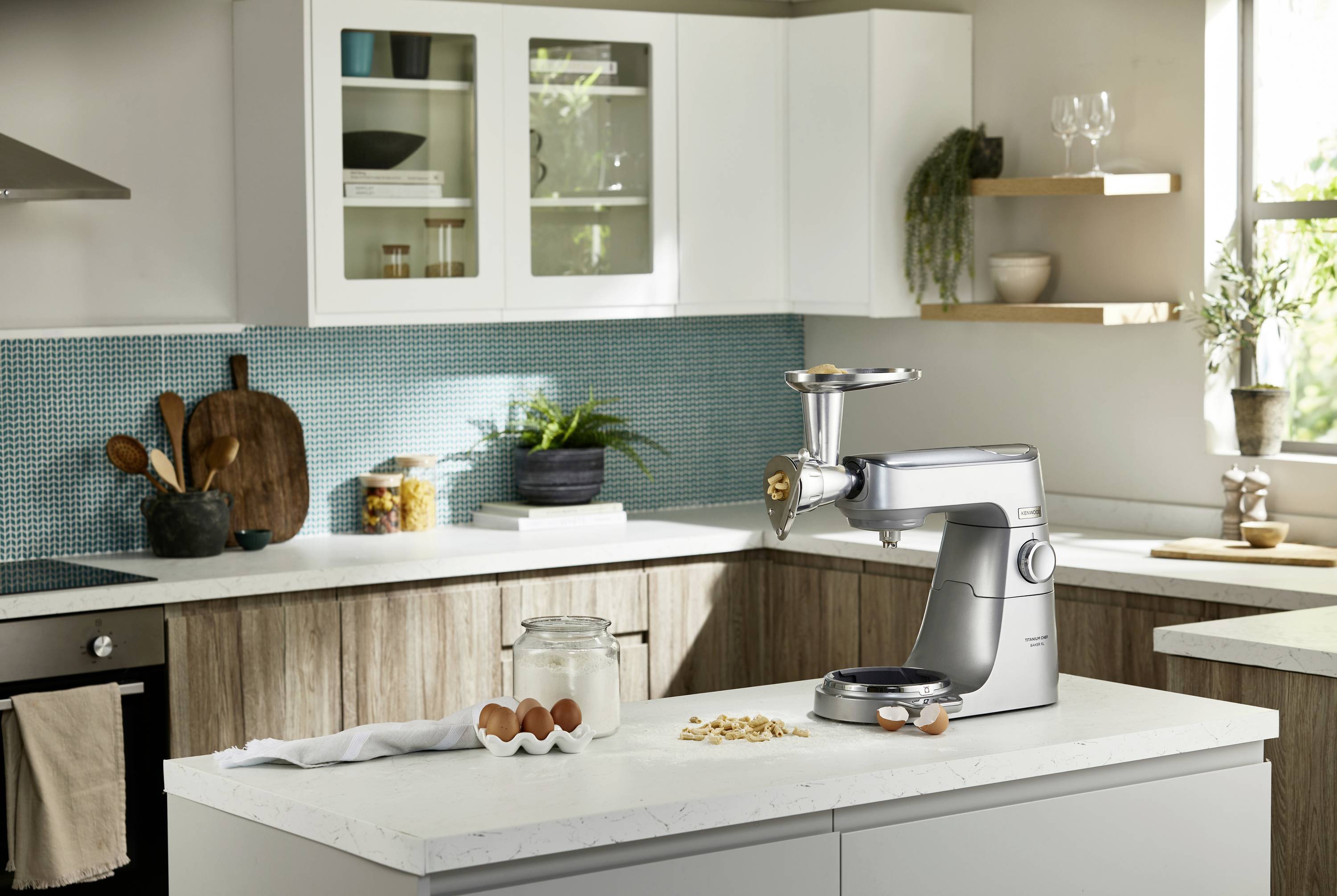 Eine moderne Küche mit weißen Schränken zeigt einen Standmixer mit einem Fleischwolfaufsatz auf der Arbeitsplatte, umgeben von Eiern und Mehl.