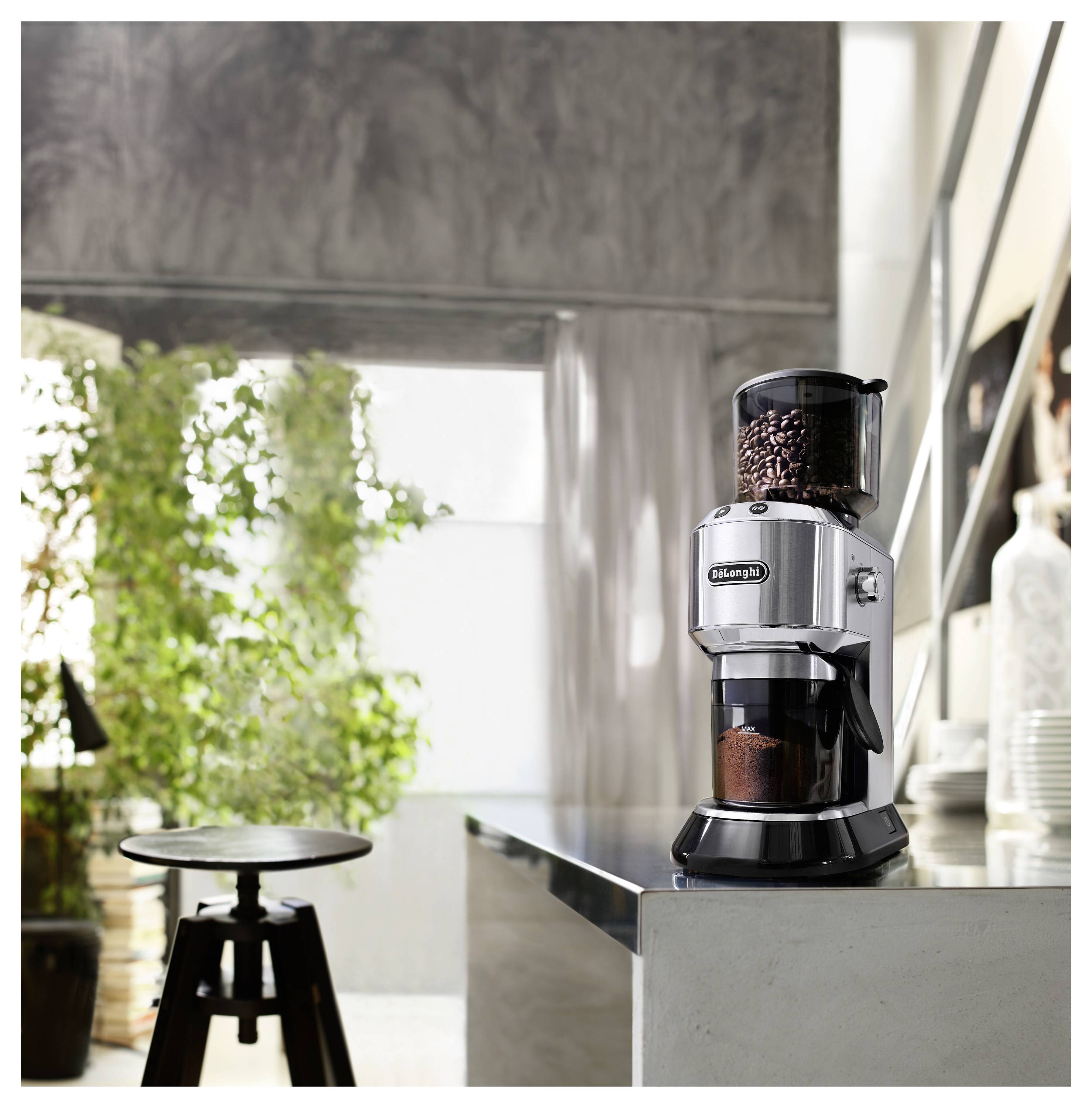 Eine silberne Kaffeemühle mit einem vollen Bohnenbehälter steht auf einer modernen Küchenarbeitsplatte, neben einer Pflanze und einem leeren Hocker.