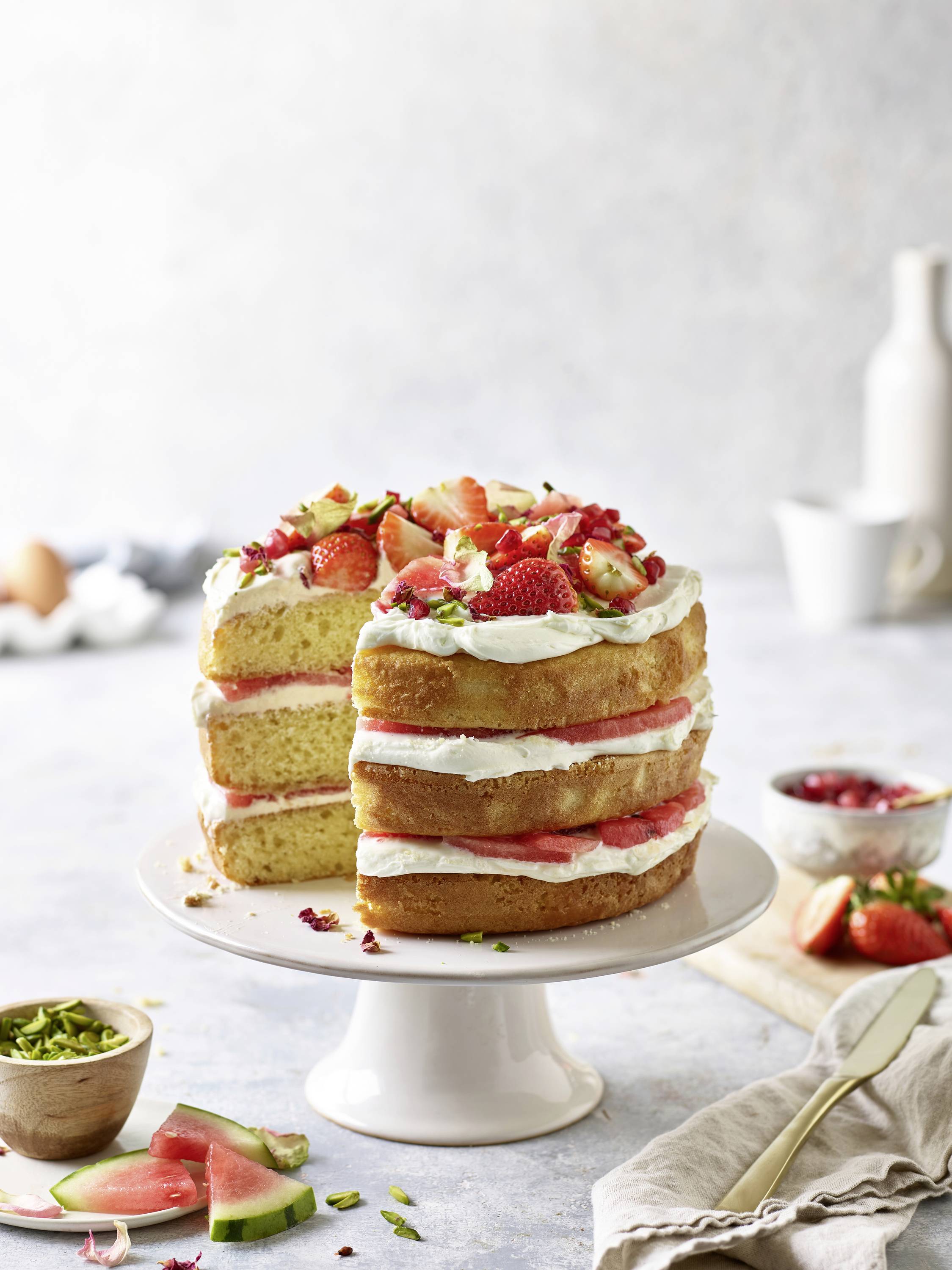 Eine geschichtete Vanilletorte mit Schlagsahne und frischen Erdbeeren, platziert auf einem weißen Kuchenständer. Eine Gabel liegt auf einem Serviettchen daneben.