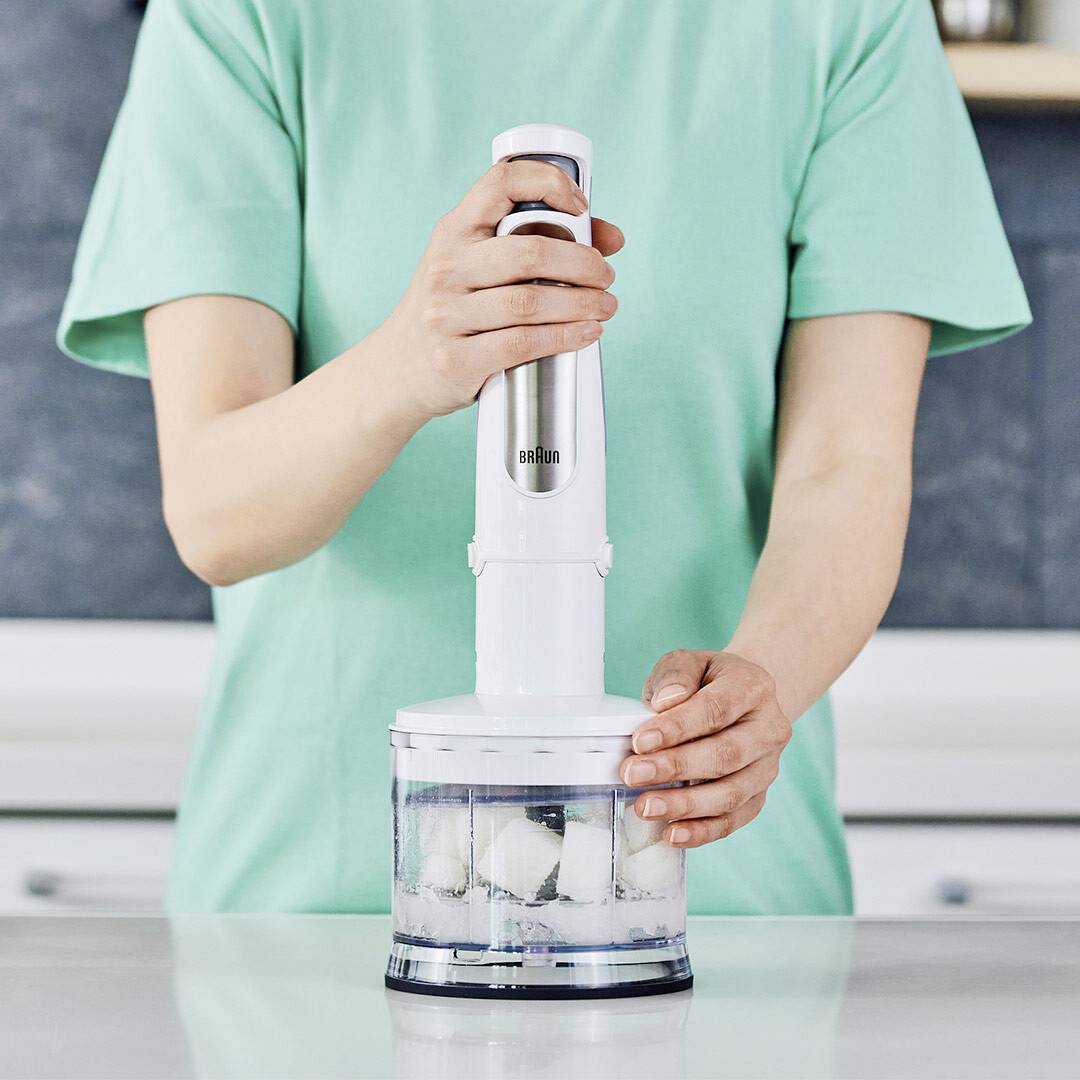 Eine Person, die einen Stabmixer verwendet, um Eiswürfel in einer Küchenumgebung zu zerkleinern, trägt ein hellgrünes Hemd.