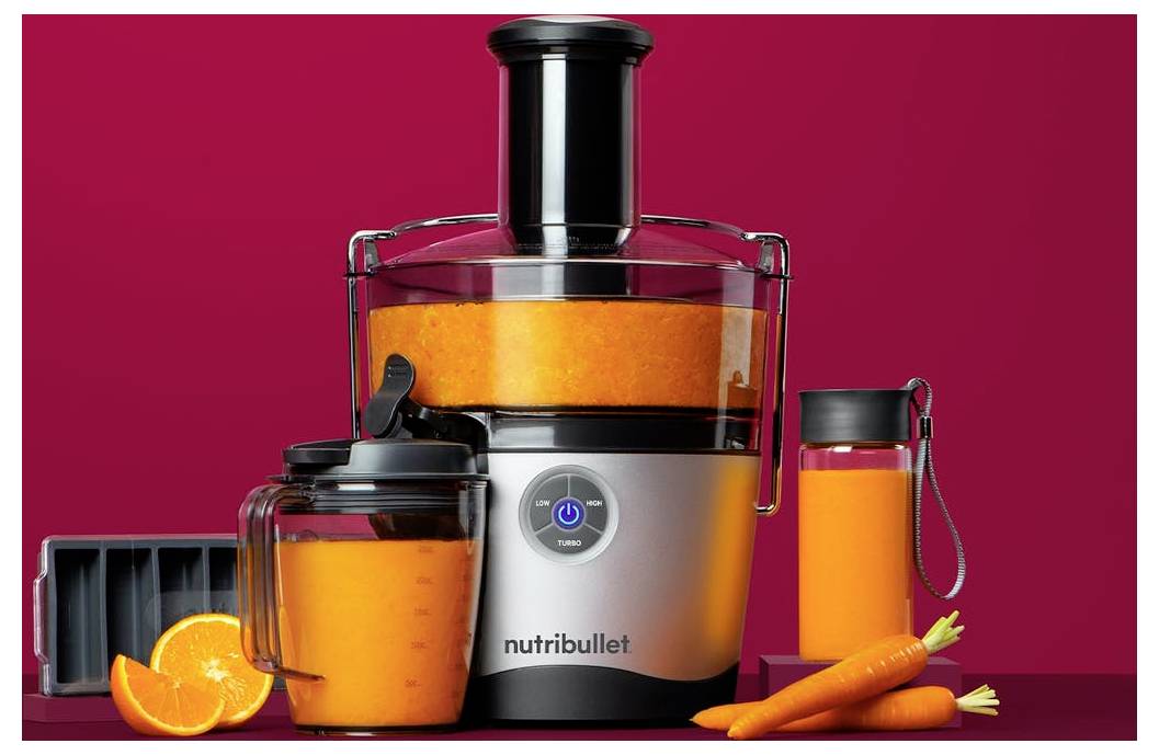 Ein silberner und schwarzer Nutribullet-Entsafter steht auf einem roten Hintergrund, umgeben von einem durchsichtigen Krug Orangensaft, einer Saftflasche, Karotten und einer Orangenscheibe.