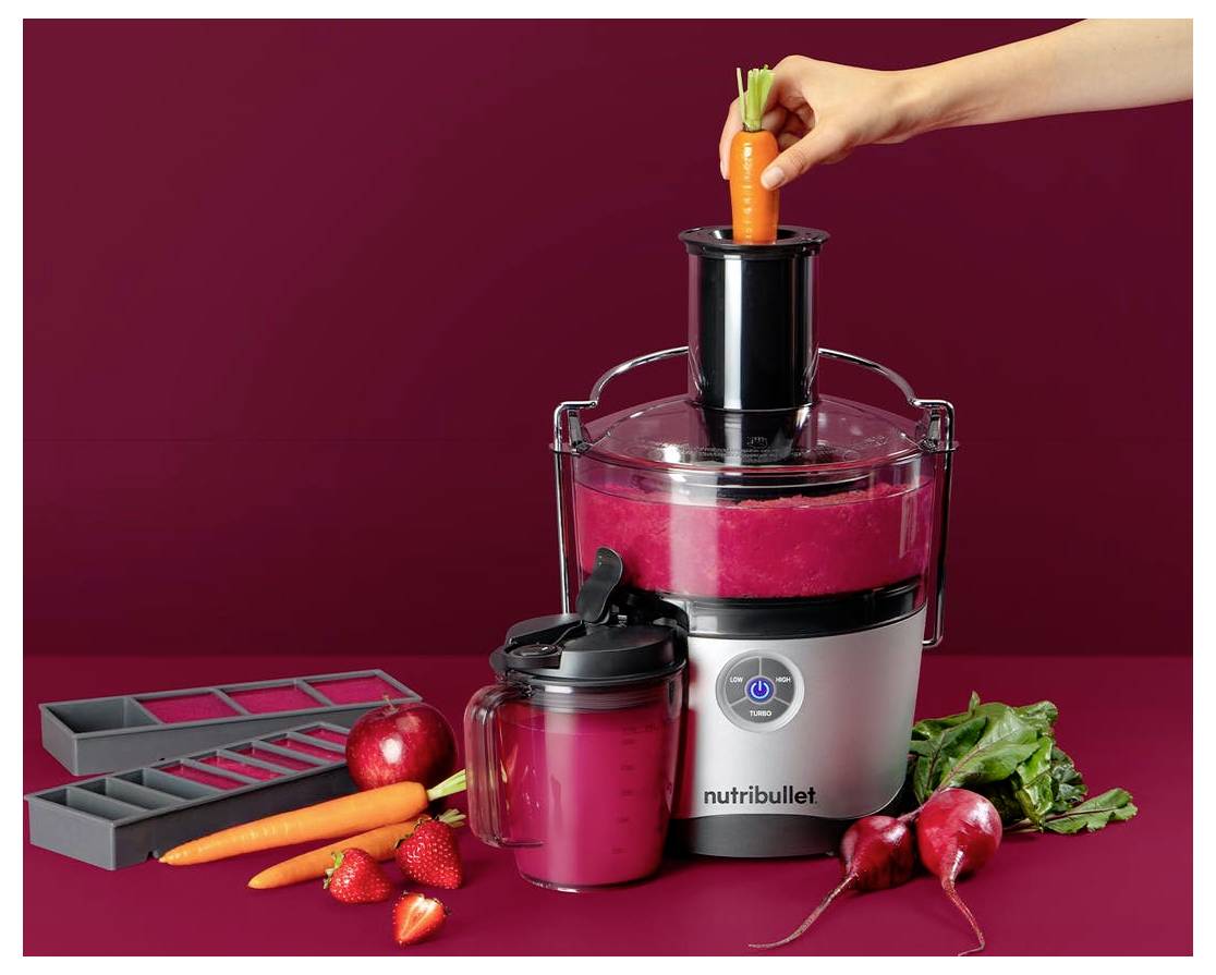 Eine Hand schiebt eine Karotte in einen Entsafter mit der Aufschrift "Nutribullet", umgeben von frischem Obst und Gemüse, darunter Karotten, Rote Beete und Erdbeeren.