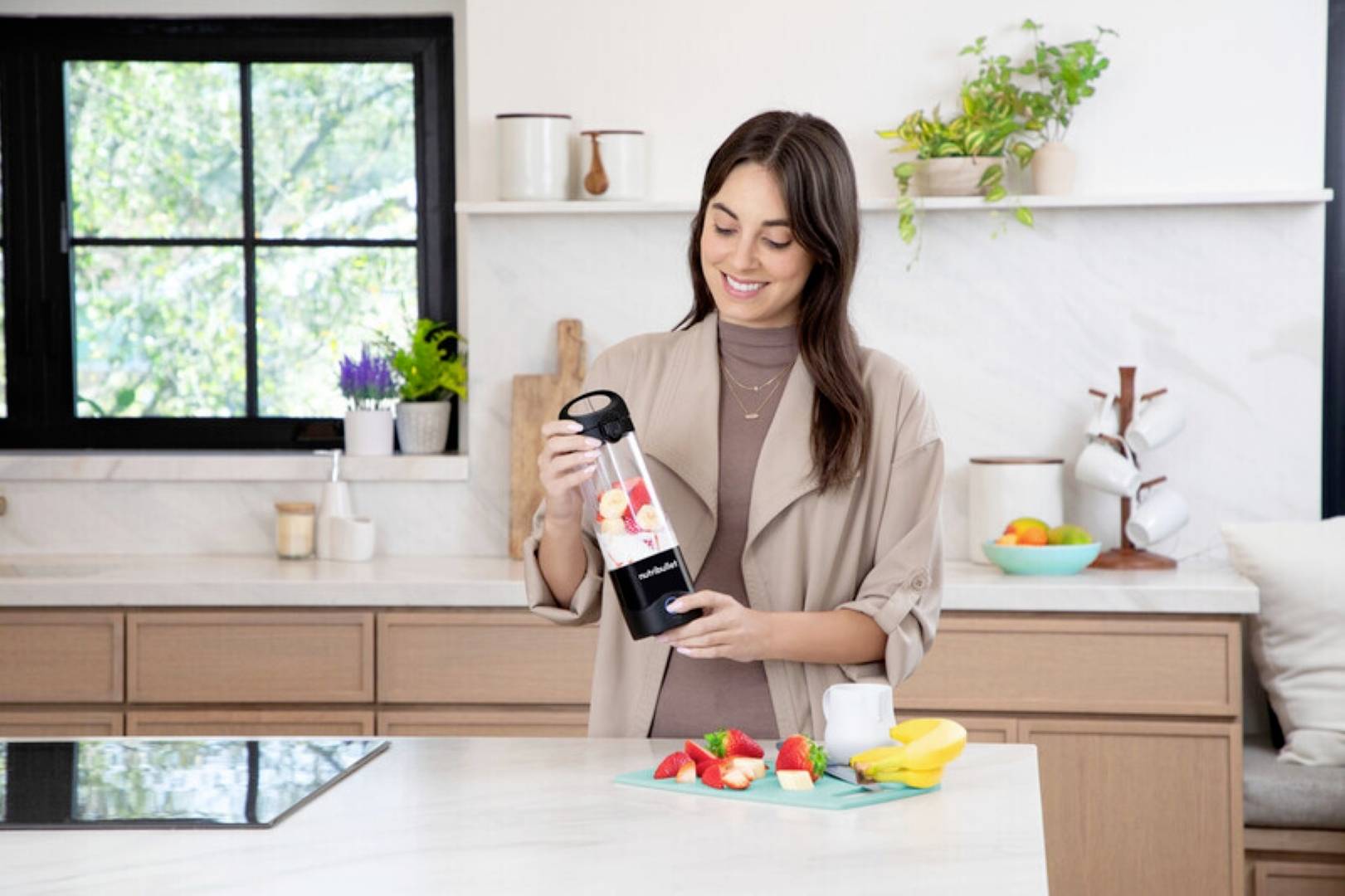 Eine Frau in einer Küche gibt geschnittene Früchte in einen Mixer. In der Nähe befinden sich weitere Früchte und ein Schneidebrett. Ein Fenster und Regale sind im Hintergrund zu sehen.
