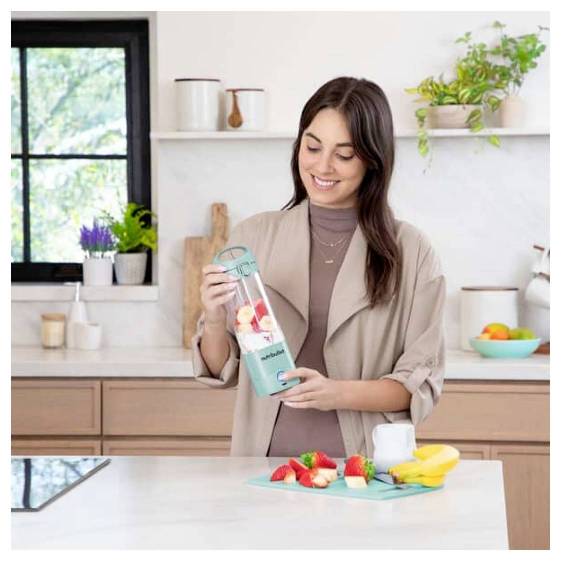 Eine Frau in einer Küche bereitet einen Smoothie mit einem Mixer zu. In der Nähe liegen Bananen und Erdbeeren auf einem Schneidebrett. Die Küche ist hell und modern.