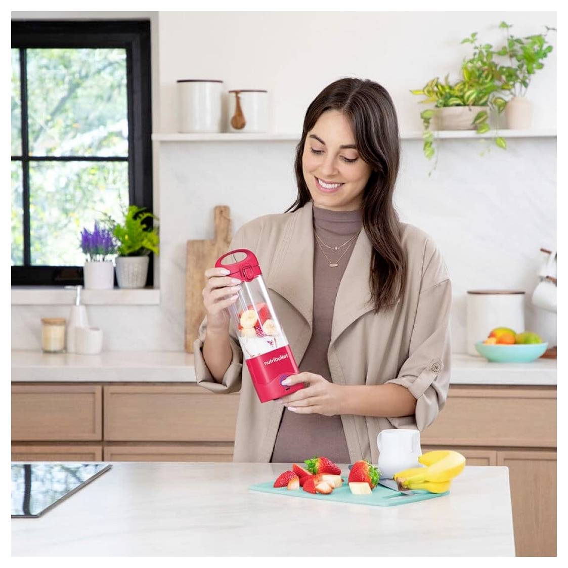 Eine Person in einer Küche, die einen kompakten Mixer mit Früchten hält, mit Fruchtstücken und einer Pflanze im Hintergrund.