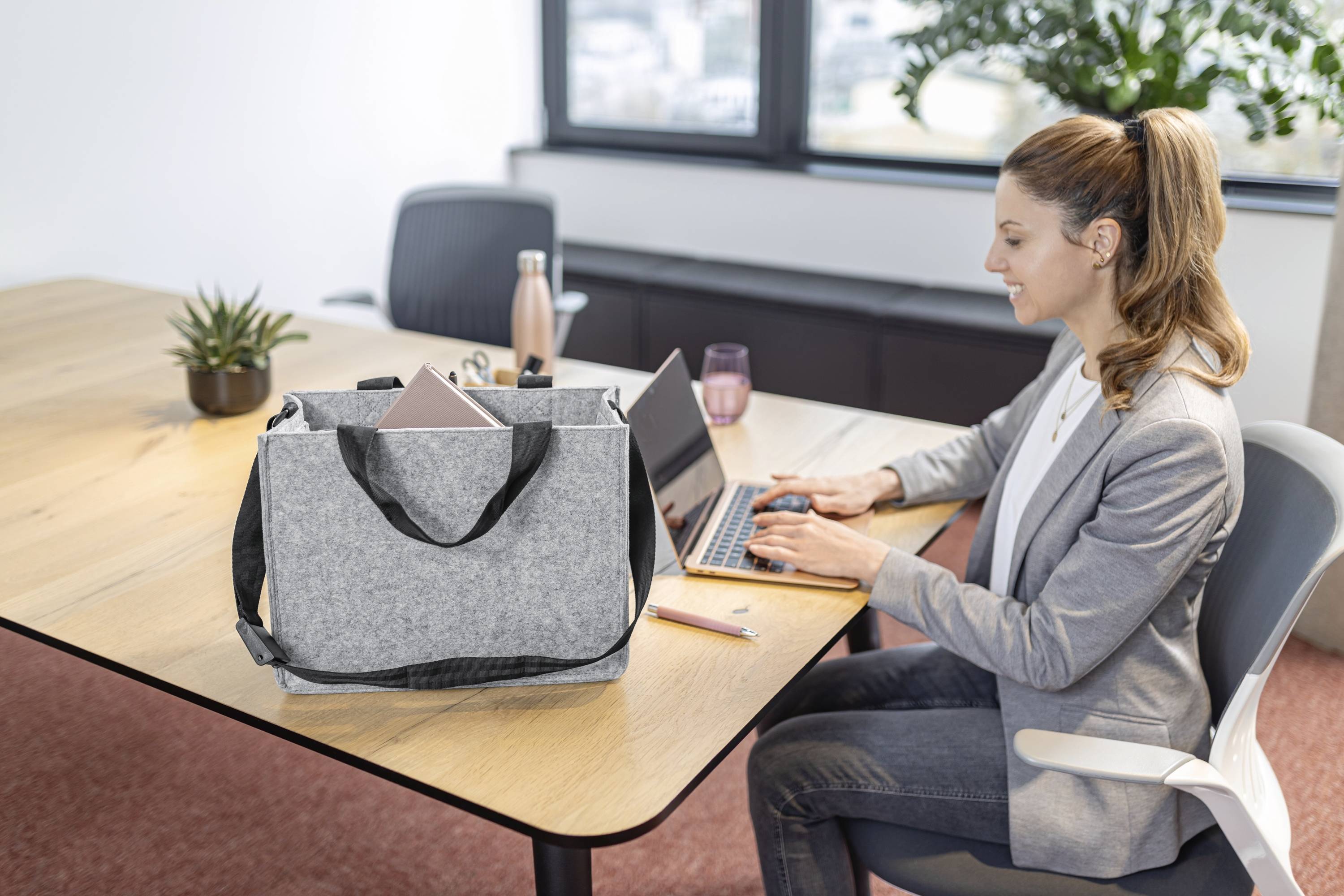 Eine Frau in einem grauen Blazer arbeitet an einem Laptop an einem Bürotisch. Eine graue Tragetasche mit einem Laptop liegt auf dem Tisch. Eine Pflanze steht in der Nähe.