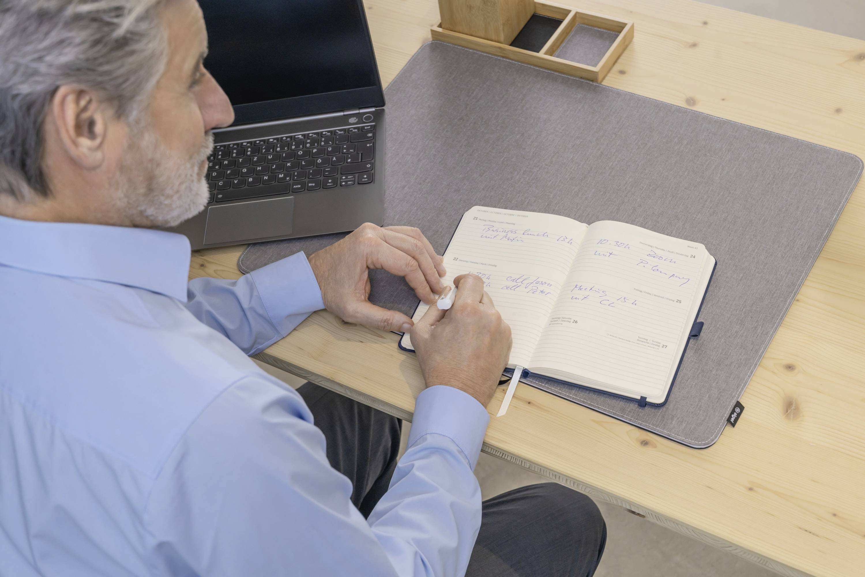 Eine Person in einem blauen Hemd schreibt in ein Notizbuch, während sie an einem Schreibtisch sitzt, auf dem ein Laptop in der Nähe steht.