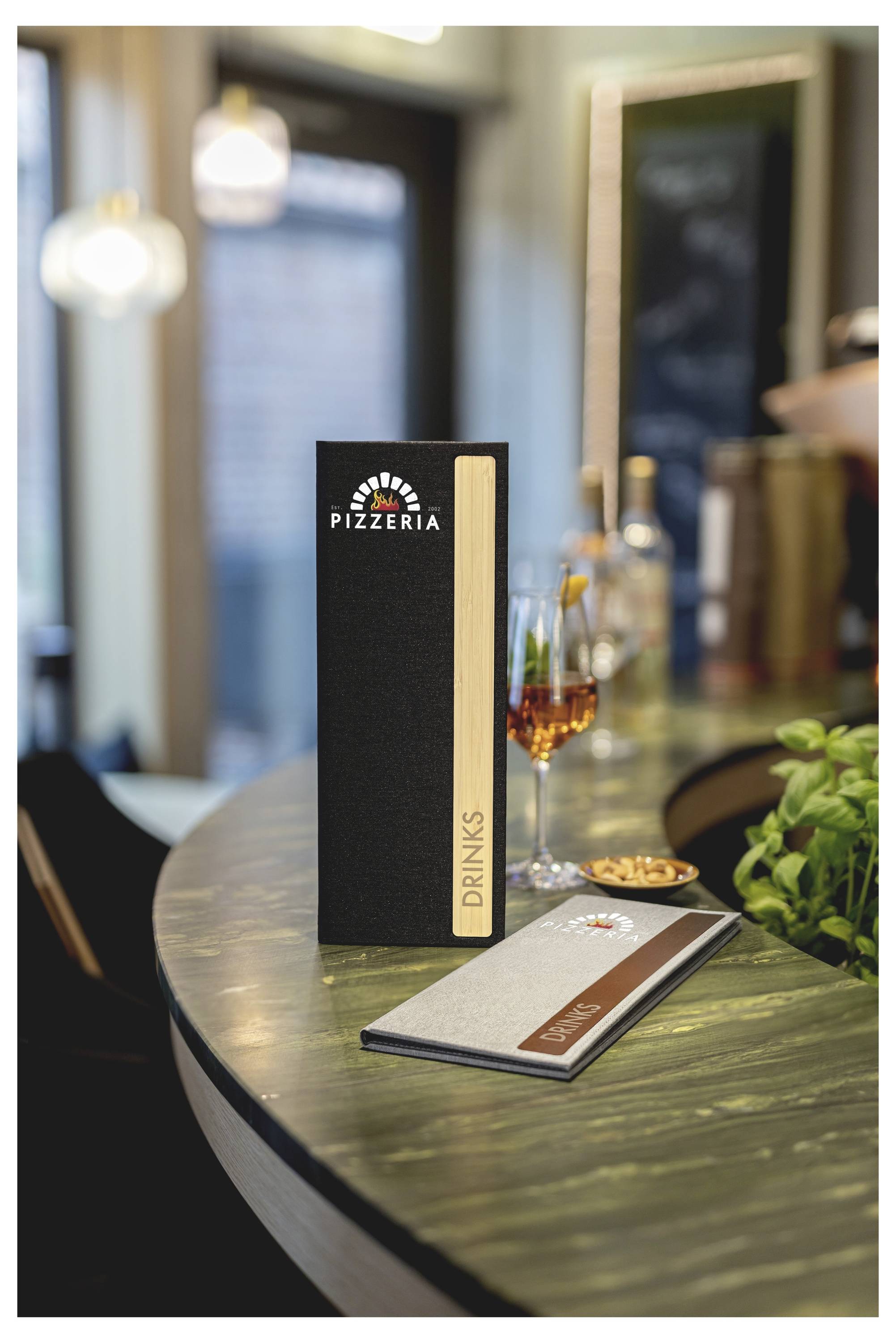 Restauranttisch mit einem schwarzen aufrecht stehenden „Pizzeria"-Menü neben einer Getränkekarte, einem Weinglas und einer Basilikumpflanze im Hintergrund.