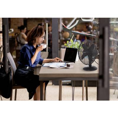 Eine Frau sitzt an einem Cafétisch, trinkt Kaffee und arbeitet an einem Laptop. Ein Ventilator und eine kleine Pflanze stehen auf dem Tisch. Drinnen, entspannte Atmosphäre.