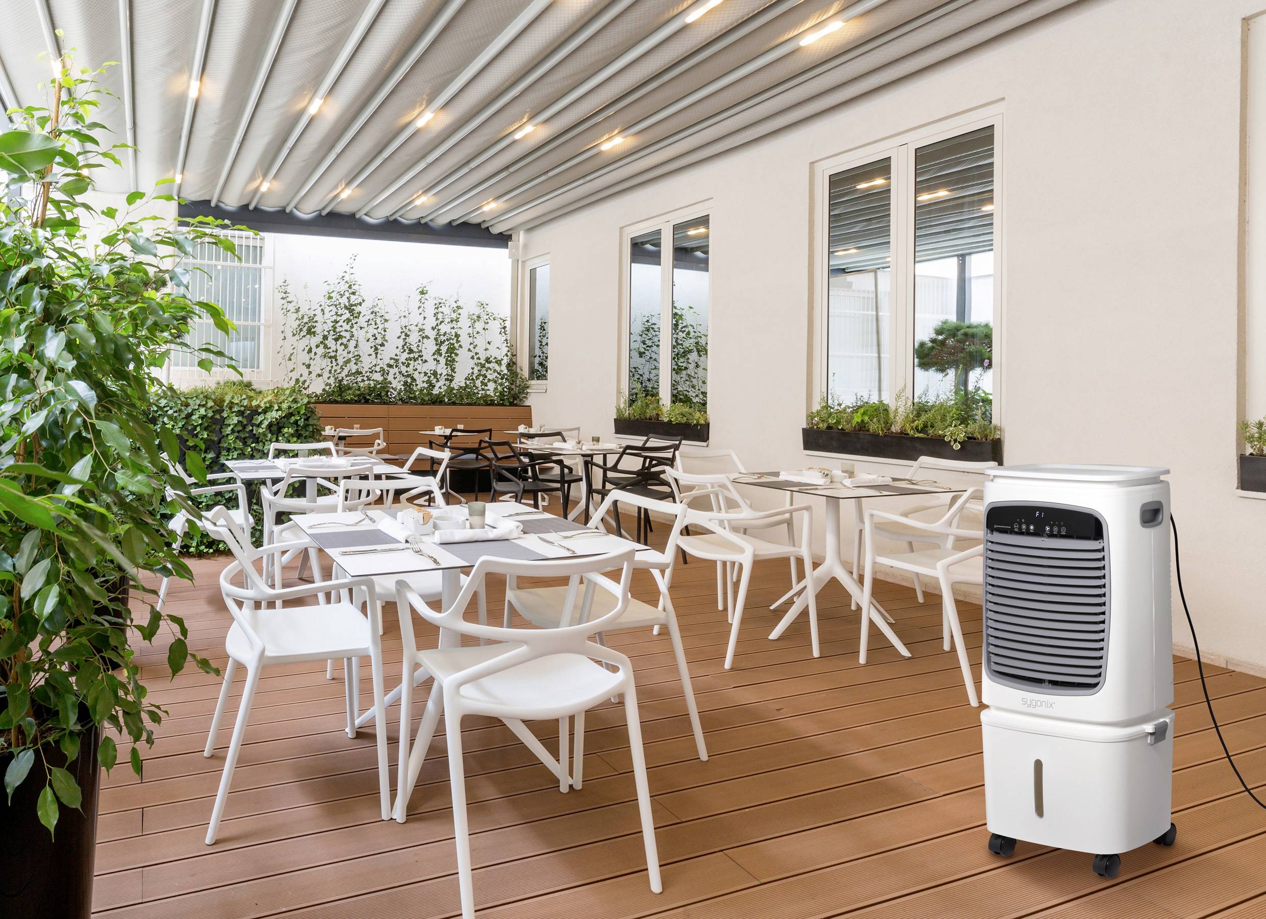 Überdachte Terrasse mit weißen Tischen und Stühlen unter einer Pergola, umgeben von Grünpflanzen. Ein mobiles Luftkühlgerät befindet sich im Vordergrund.