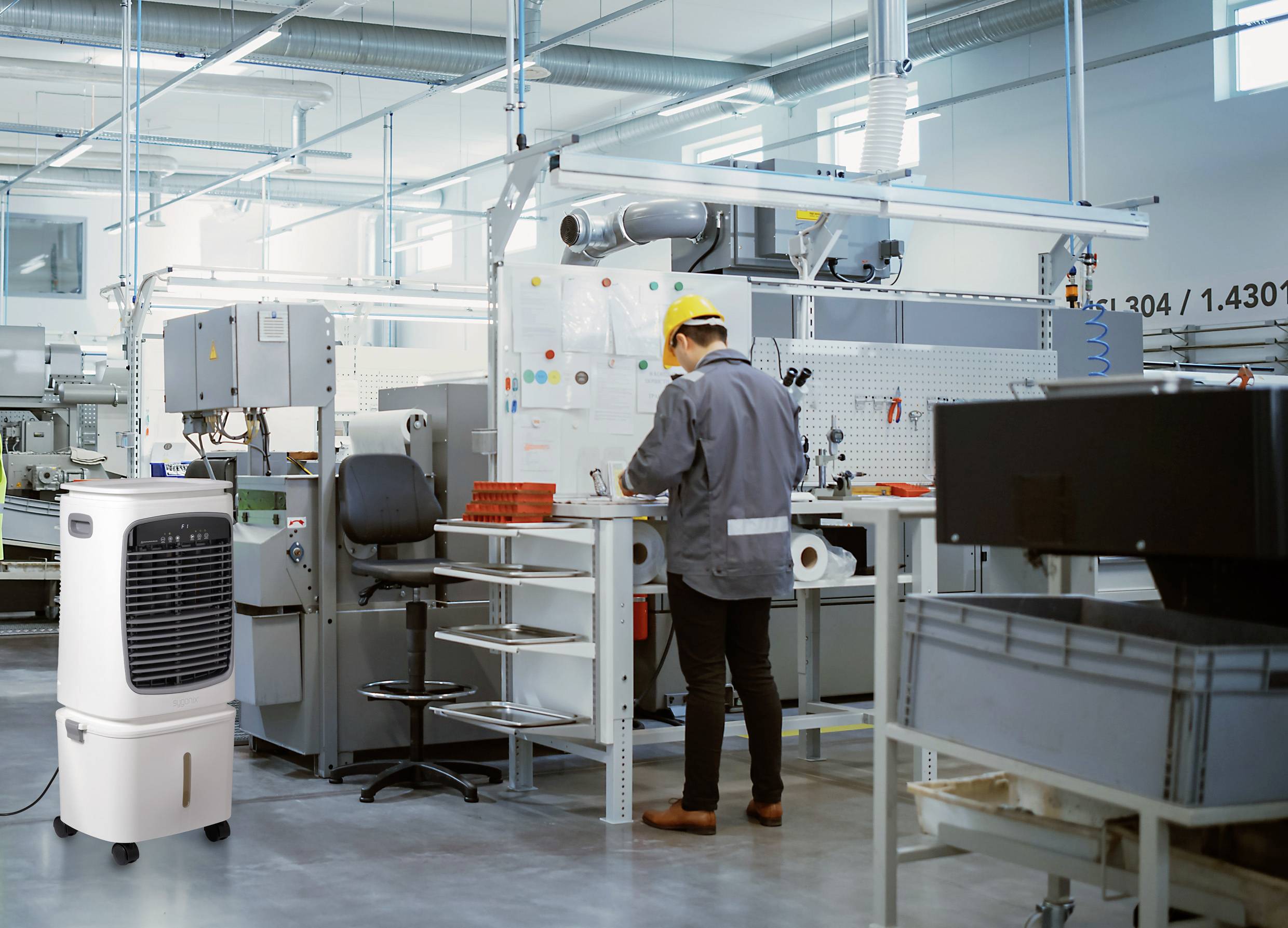 Ein Fabrikarbeiter mit gelbem Schutzhelm kontrolliert Ausrüstung an einem Bedienfeld, wobei industrielle Maschinen und ein mobiler Luftkühler in der Nähe stehen.