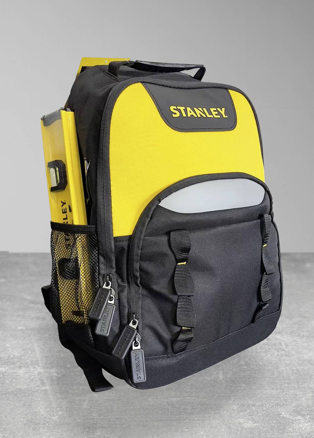 Ein schwarzer und gelber Rucksack mit der Aufschrift 'STANLEY' auf der Vorderseite, ausgestattet mit seitlichen Netztaschen und mehreren Fächern, der auf einem grauen Boden steht.