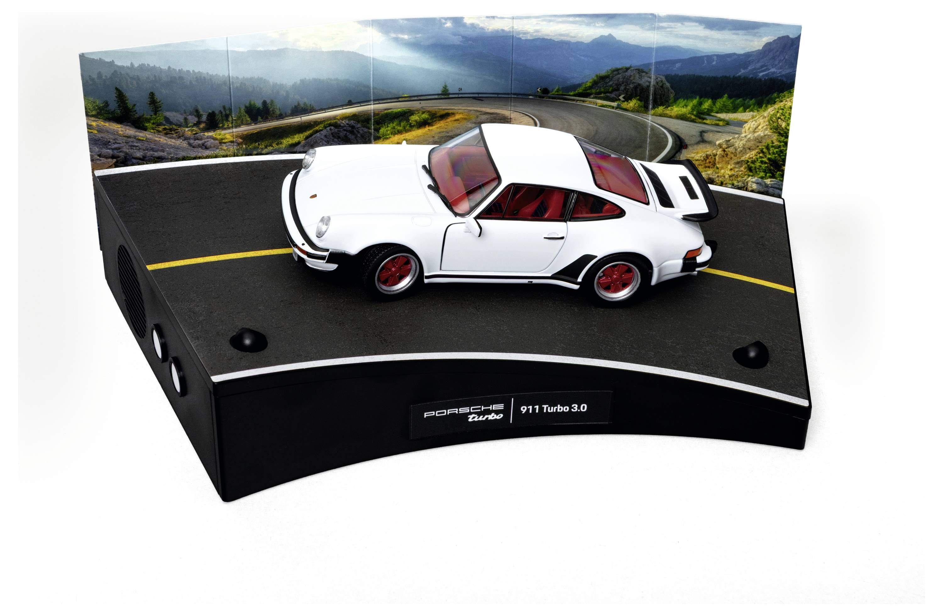 Ein weißes Modellauto mit roten Akzenten wird vor einer geschwungenen Straßenkulisse präsentiert, die bergiges Gelände zeigt und mit 'Porsche 911 Turbo 3.0' beschriftet ist.