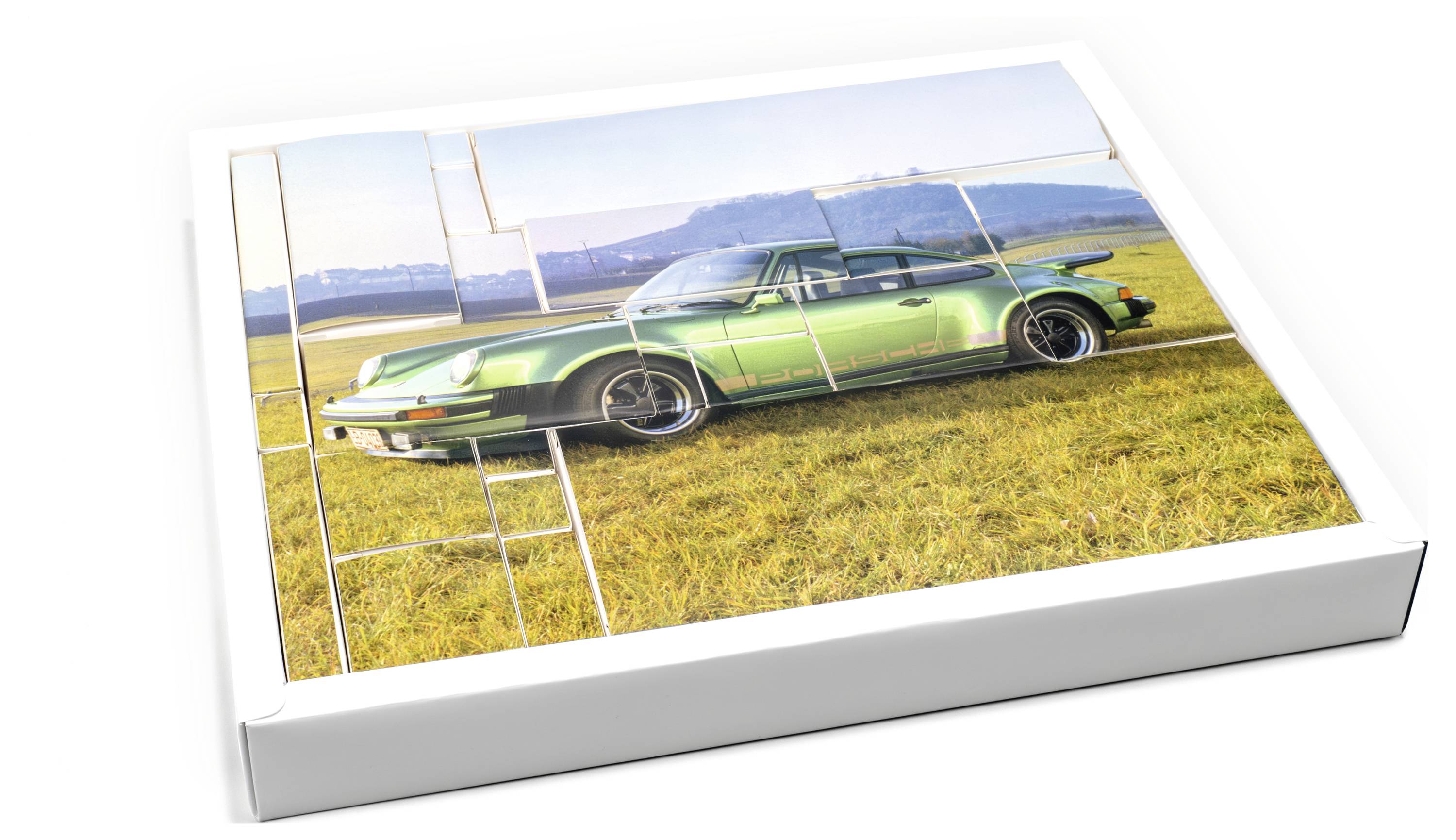 Puzzle, das einen Oldtimer-Sportwagen in Grün auf einem Feld mit Gras und Bergen im Hintergrund zeigt, in einer geöffneten Box.