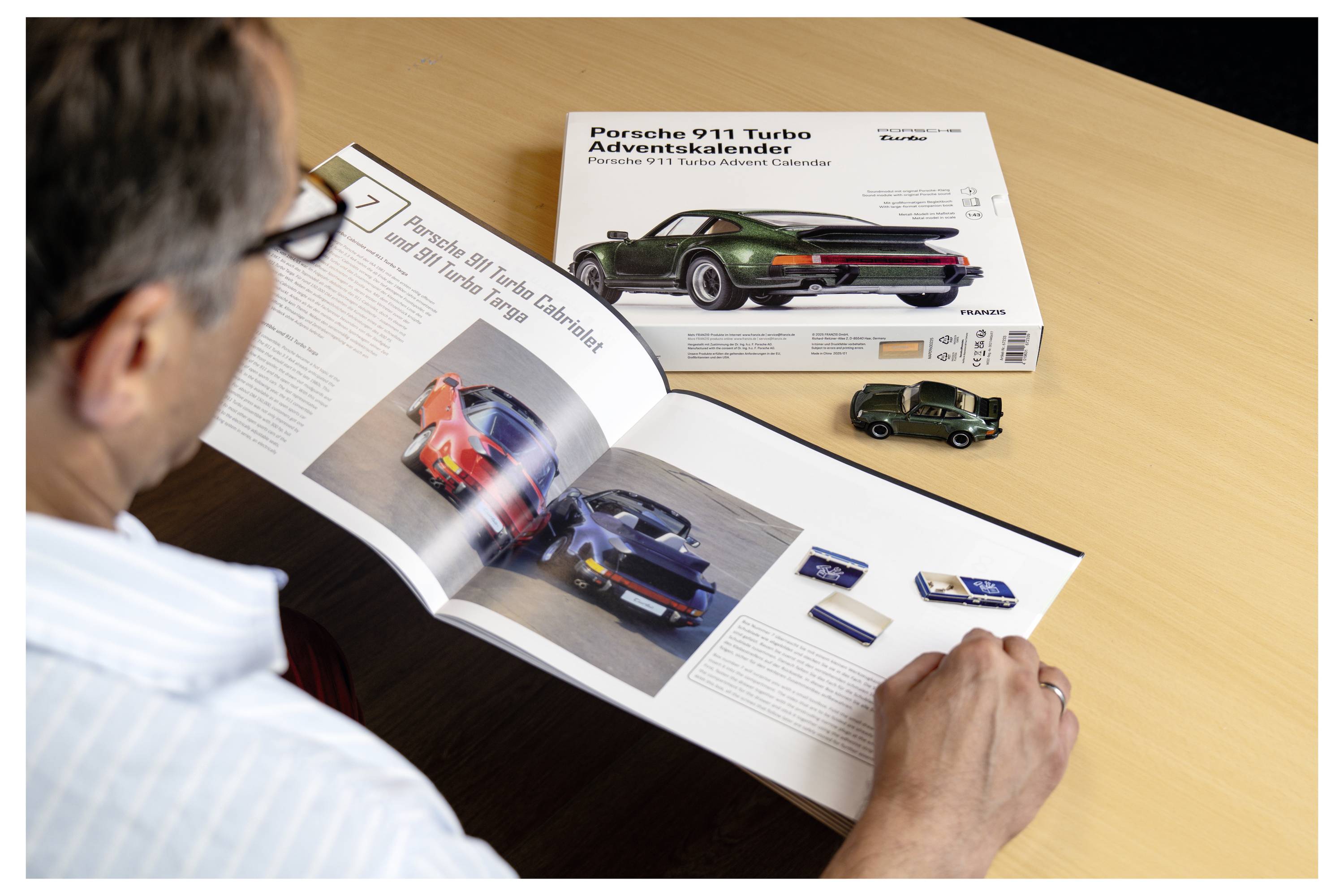 Eine Person liest ein Buch mit Autobildern neben einem Modellauto und einem Porsche 911 Turbo Adventskalender auf einem Tisch.