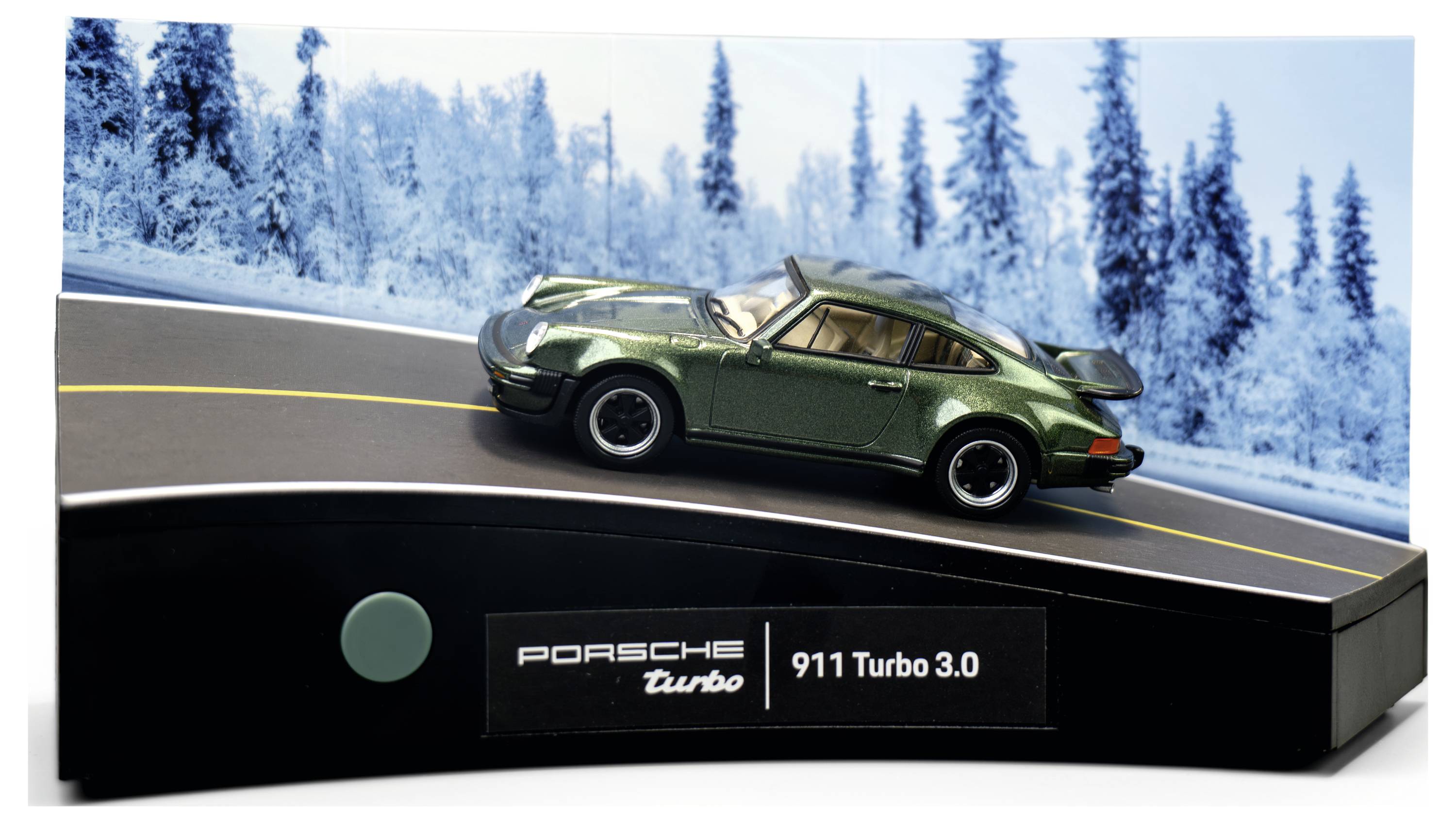 Ein grünes Modellauto mit der Aufschrift „Porsche 911 Turbo 3.0" wird auf einer geschwungenen Straße mit einer verschneiten Baumkulisse präsentiert, was einen Winterkontext andeutet.