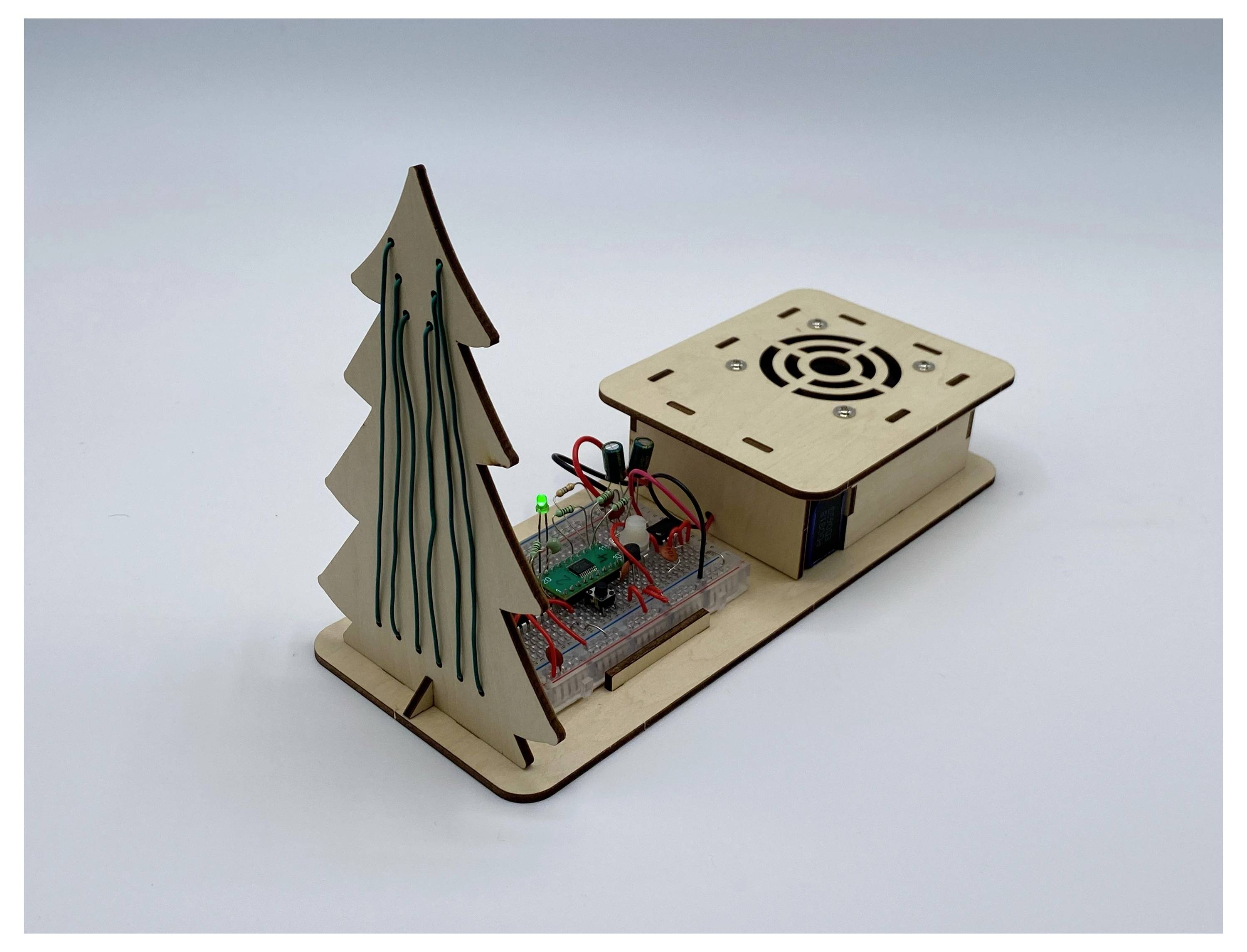 Ein dekorativer Holzbaum mit eingebauten Lichtern steht neben einer kleinen offenen Box, die elektronische Komponenten enthält, darunter eine grüne LED.