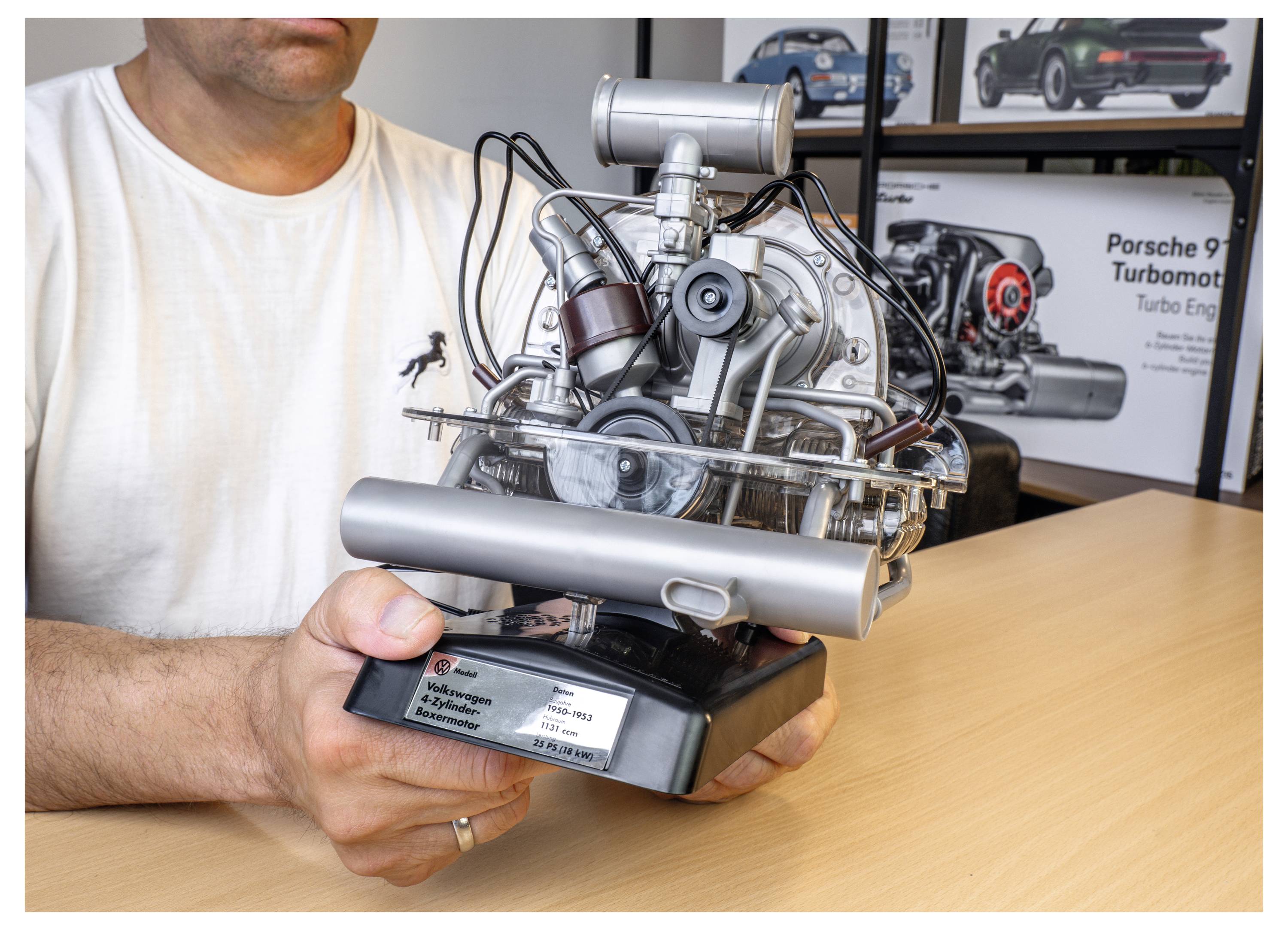 Eine Person hält ein Modell eines Porsche-911-Motors auf einem Tisch. Der Hintergrund zeigt Poster mit Motordiagrammen.